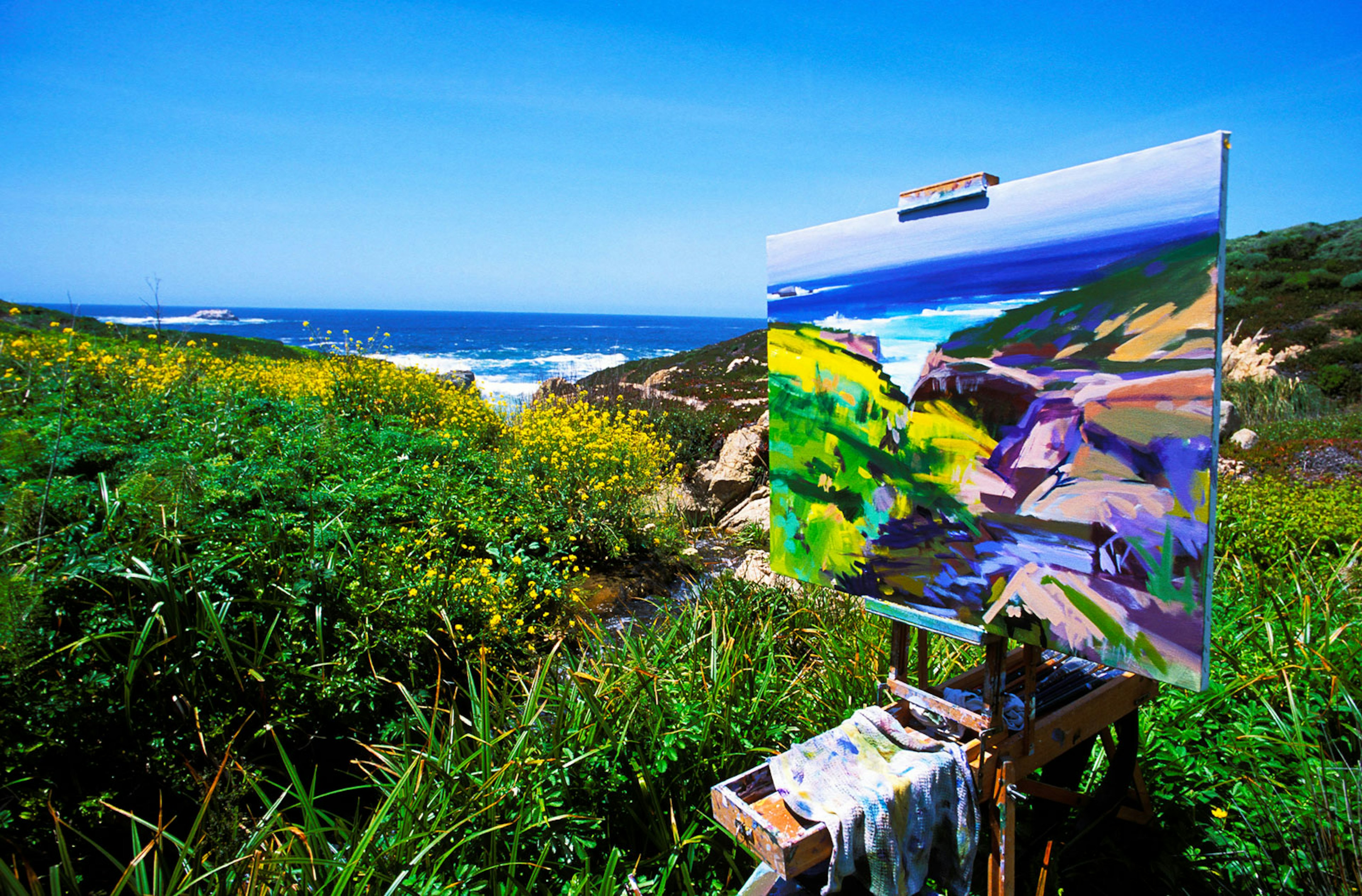 a finished landscape stands on an easel in with wildflowers and ocean in the background