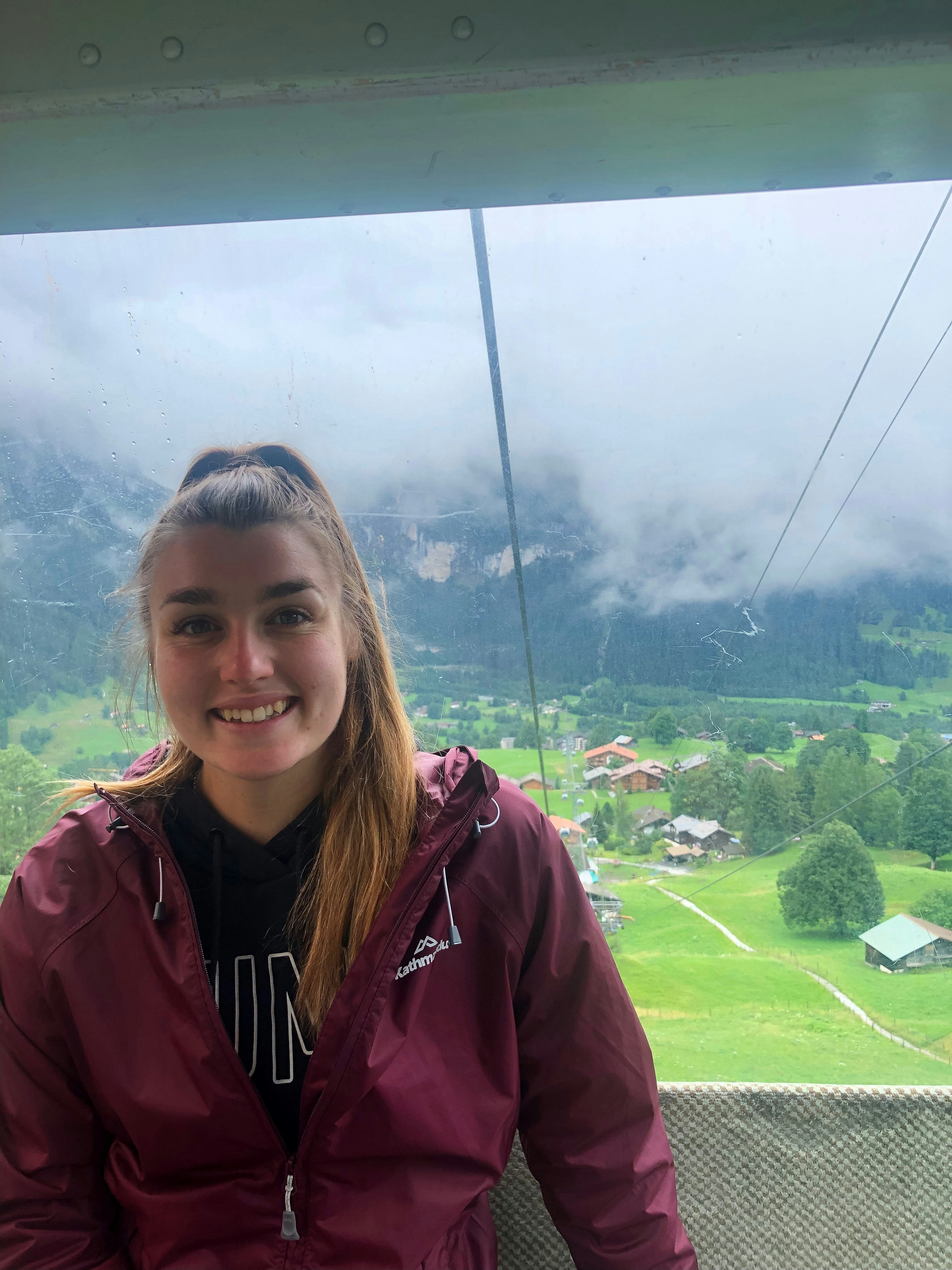 A young woman is in a cable car, riding up a mountain. She is smiling and looking at the camera and, behind her, clouds can be seen lingering above a mountain village.