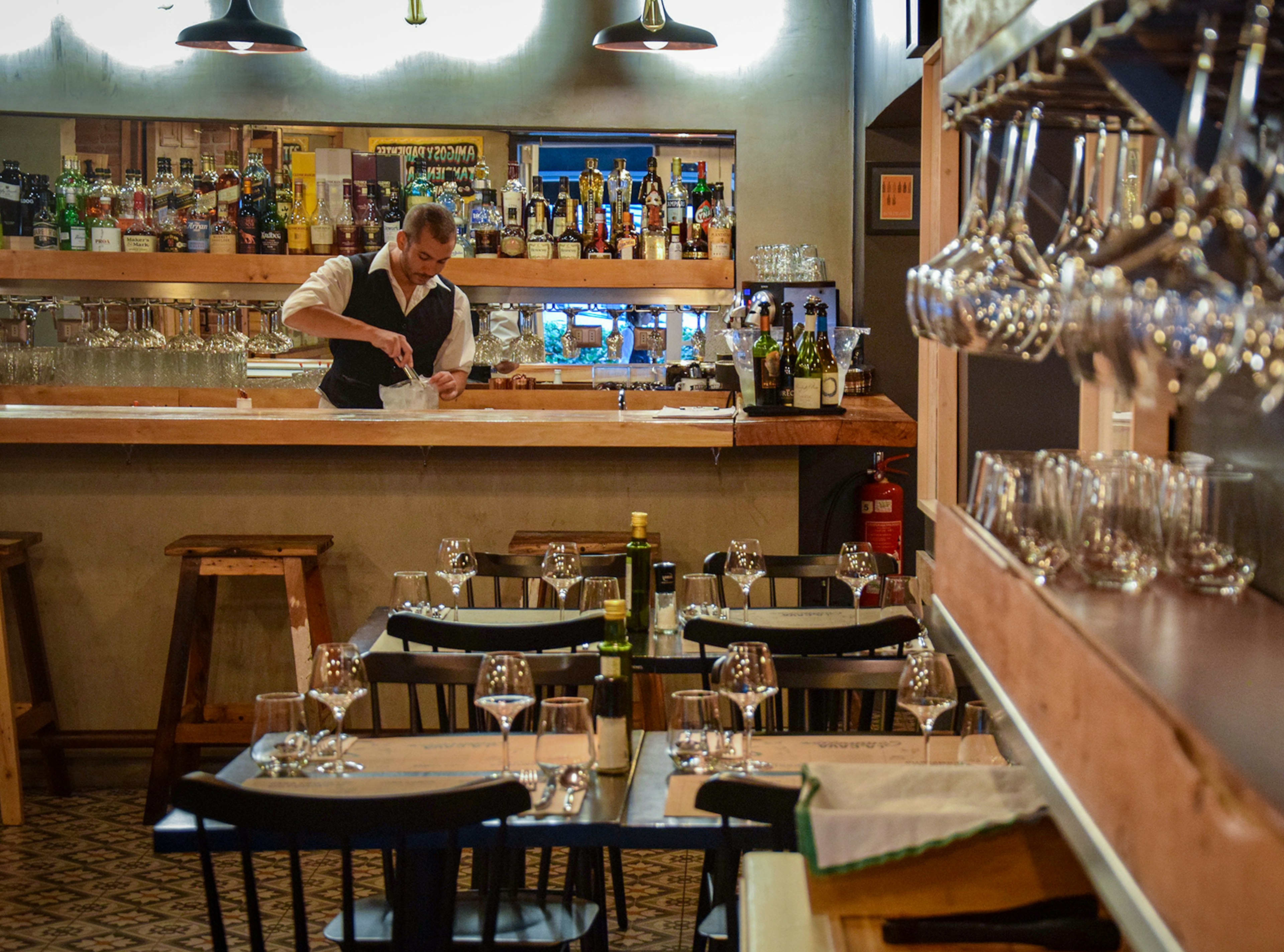 A shot of a bartender working at the bar with tables set with wine glasses in the foreground