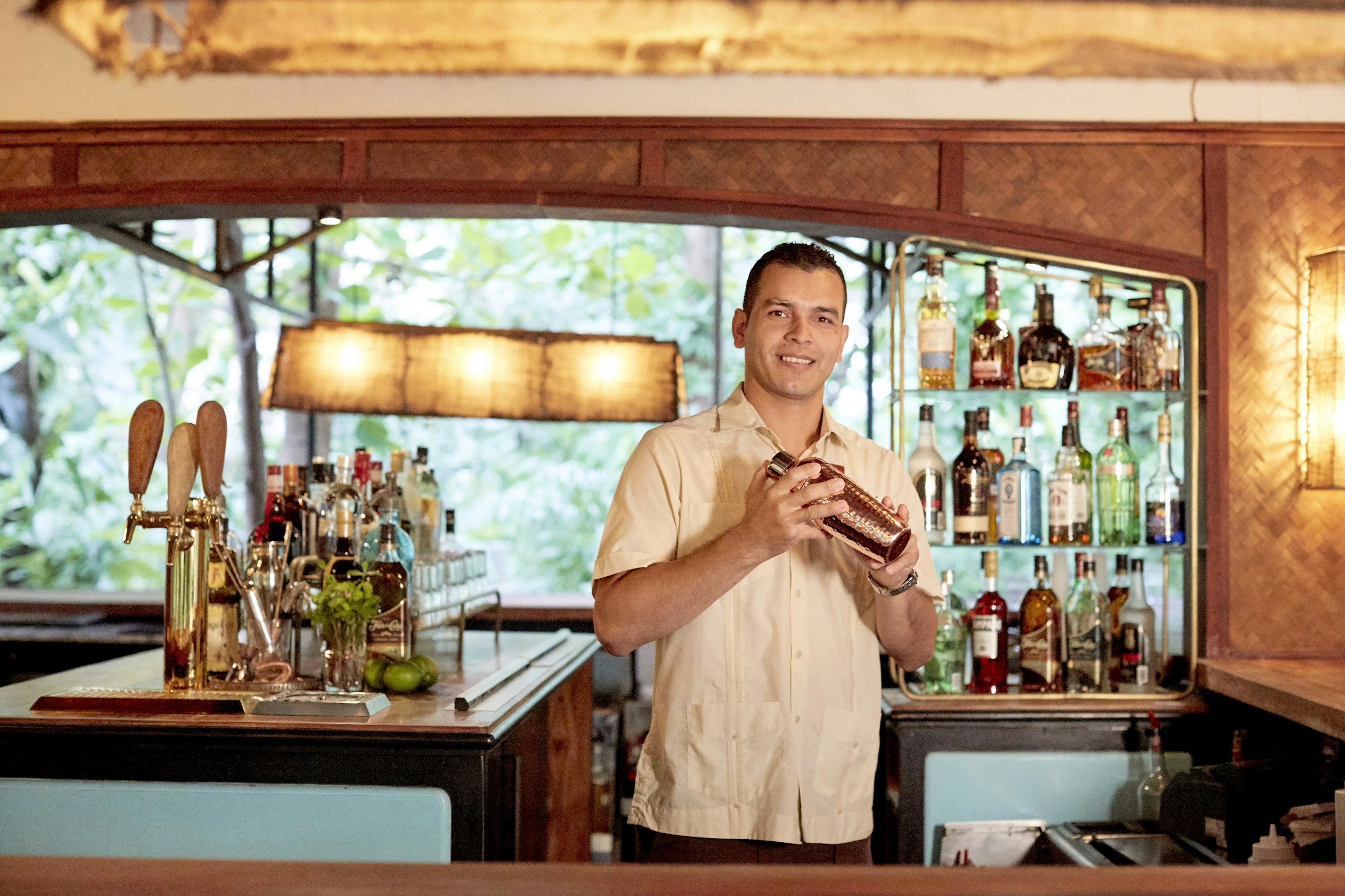 A man holding a cocktail shaker stands behind a well-stocked bar and smiles towards the camera