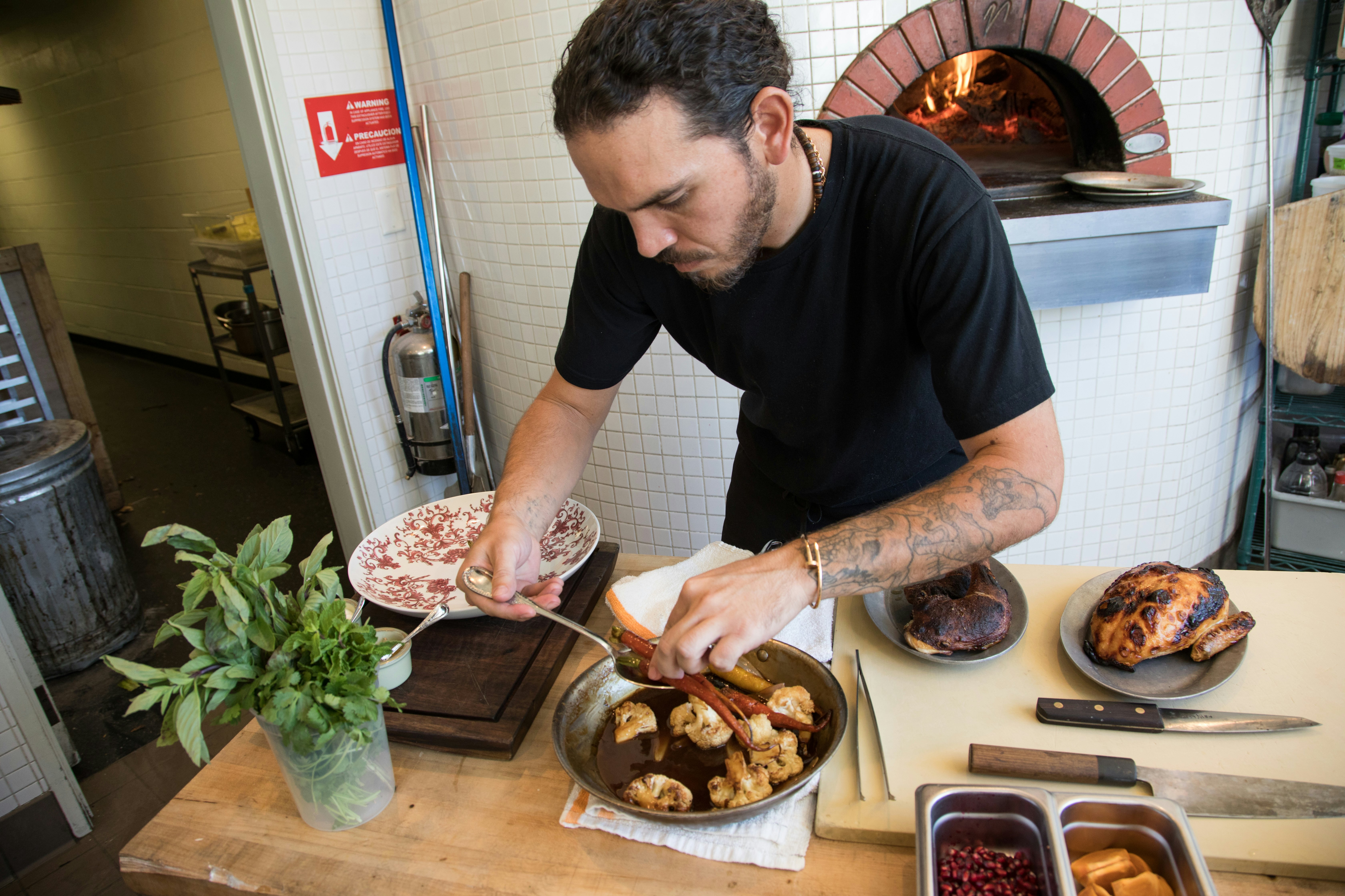 Chef Casey Lane uses a spoon to lift braised carrots out of a stir fry pan with cauliflower. There are whole chickens on separate plates on a cutting board next to him.