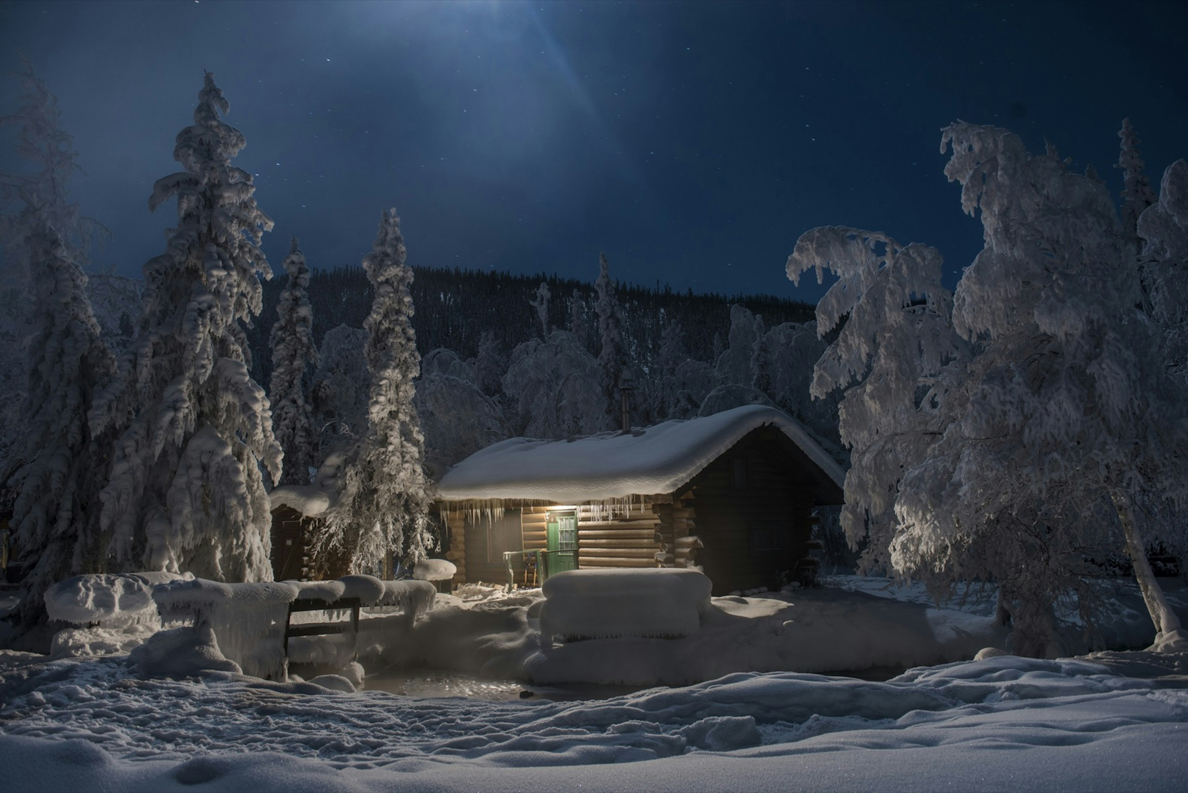 A snow-covered cabin and trees surround a water hole at night at Chena hot springs near Fairbanks Alaska.