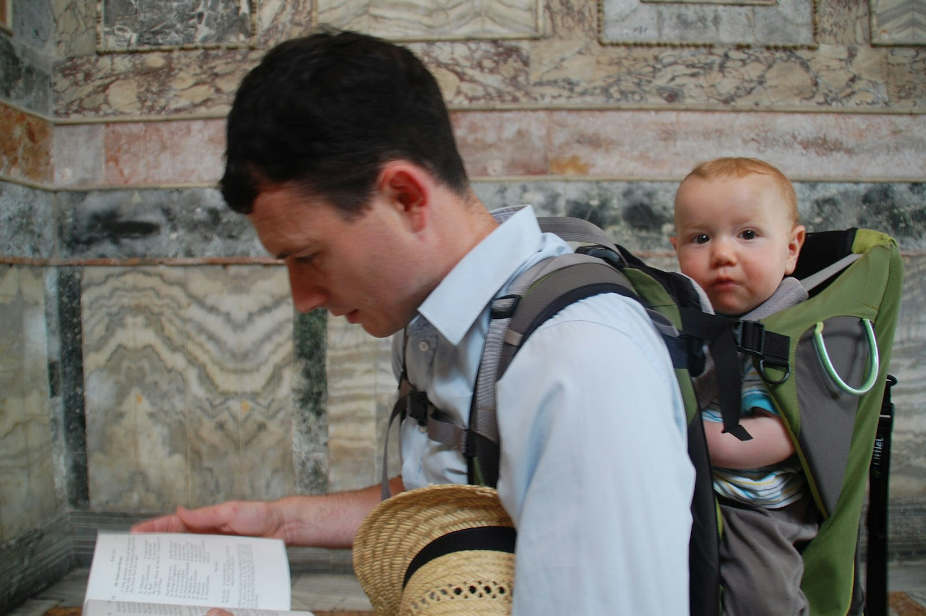 A father looks down at a guidebook within an Istanbul church while his infant child on his back looks inquisitively towards the camera