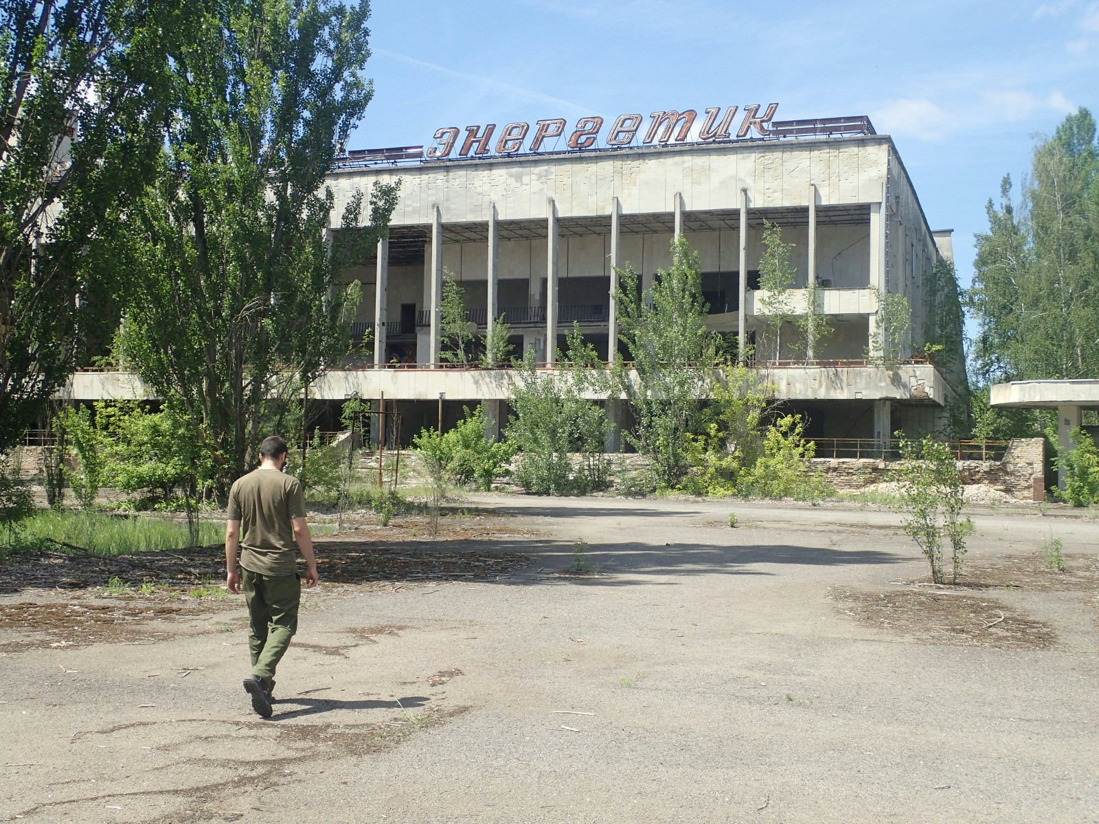 A guide walks across tarmac towards the derelict Palace of Culture in Pripyat; tress are growing in front of and within the pillared concrete structure, which has a large sign in Ukrainian script on its roof.