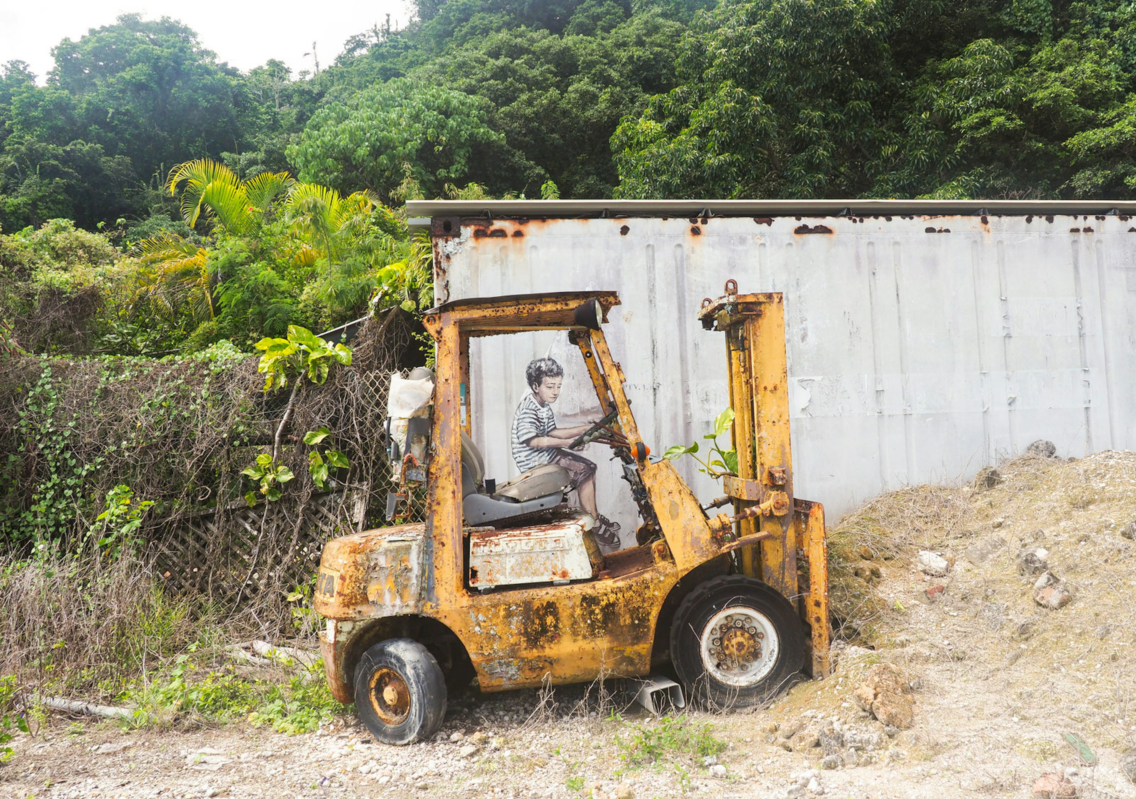 A forlorn forklift is parked next to an old shipping container with an image of a boy painted to look as if he is driving the forklift