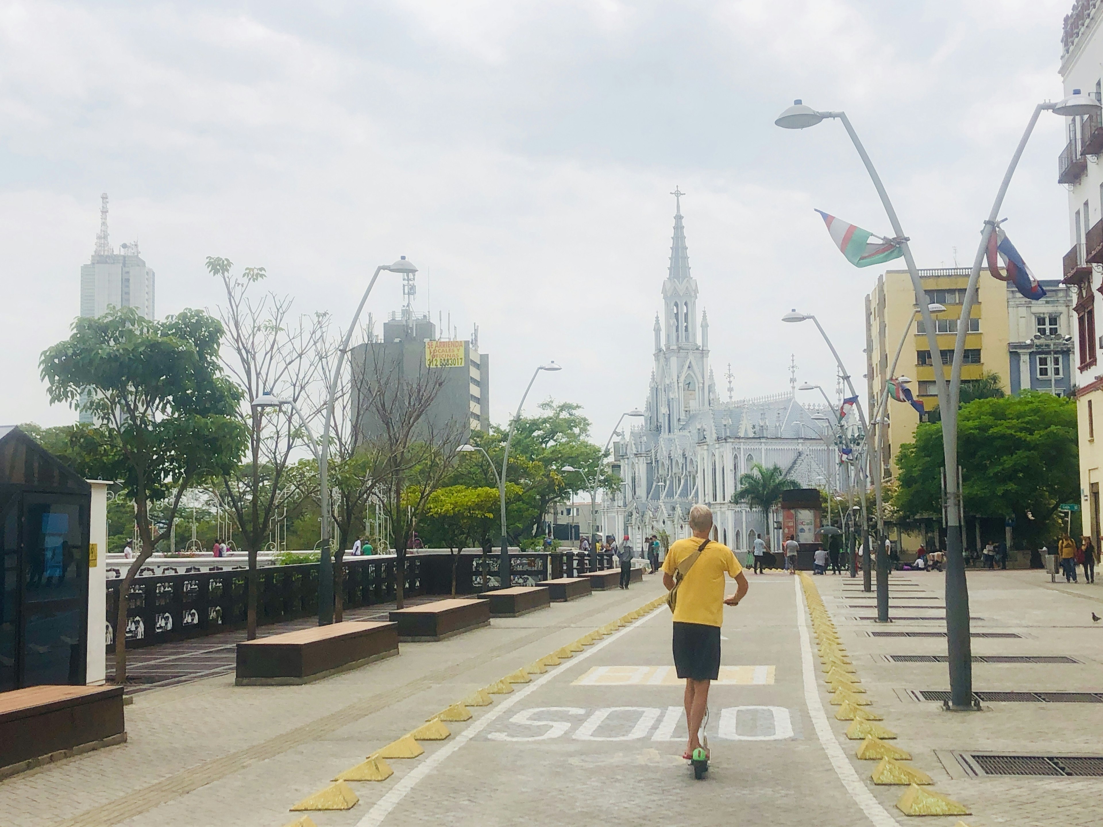 A man in a yellow shirt rides a scooter down a city bike lane towards a section of Cali;