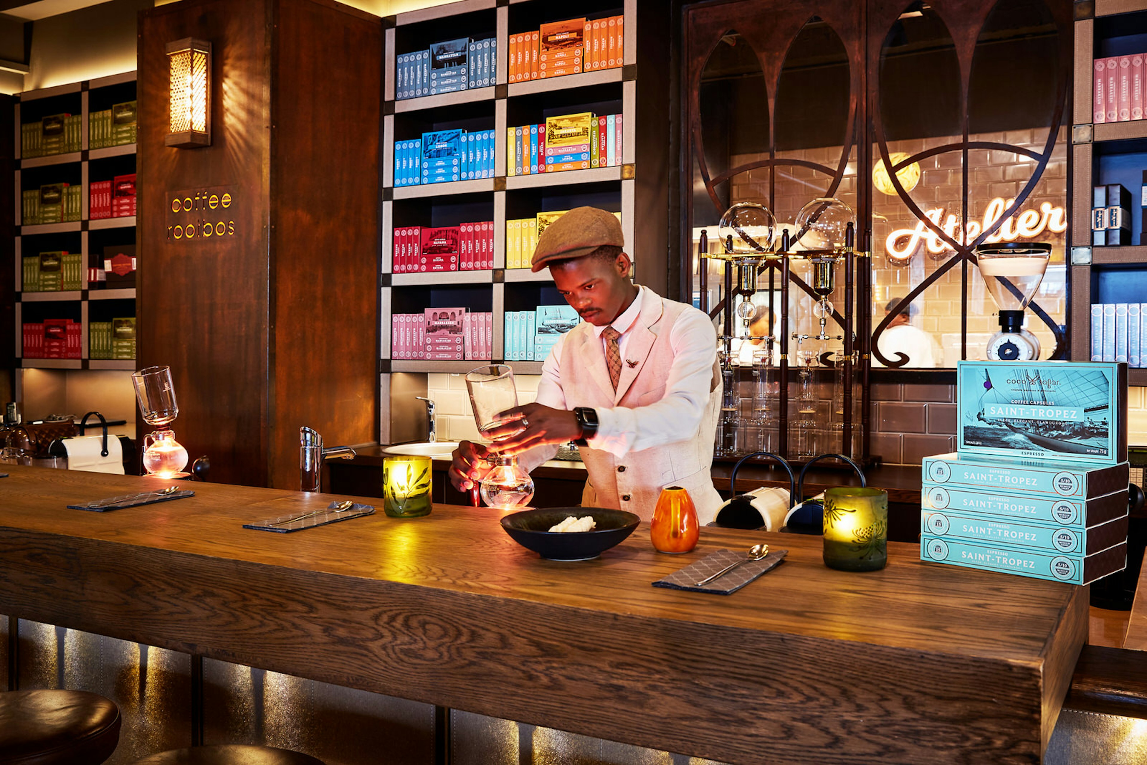 A smartly dressed barista in a flat cap, waist coat and tie concocts a coffee; the rich wood interior includes numerous shelves full of boxed coffee (each shelve holding matching coloured boxes)