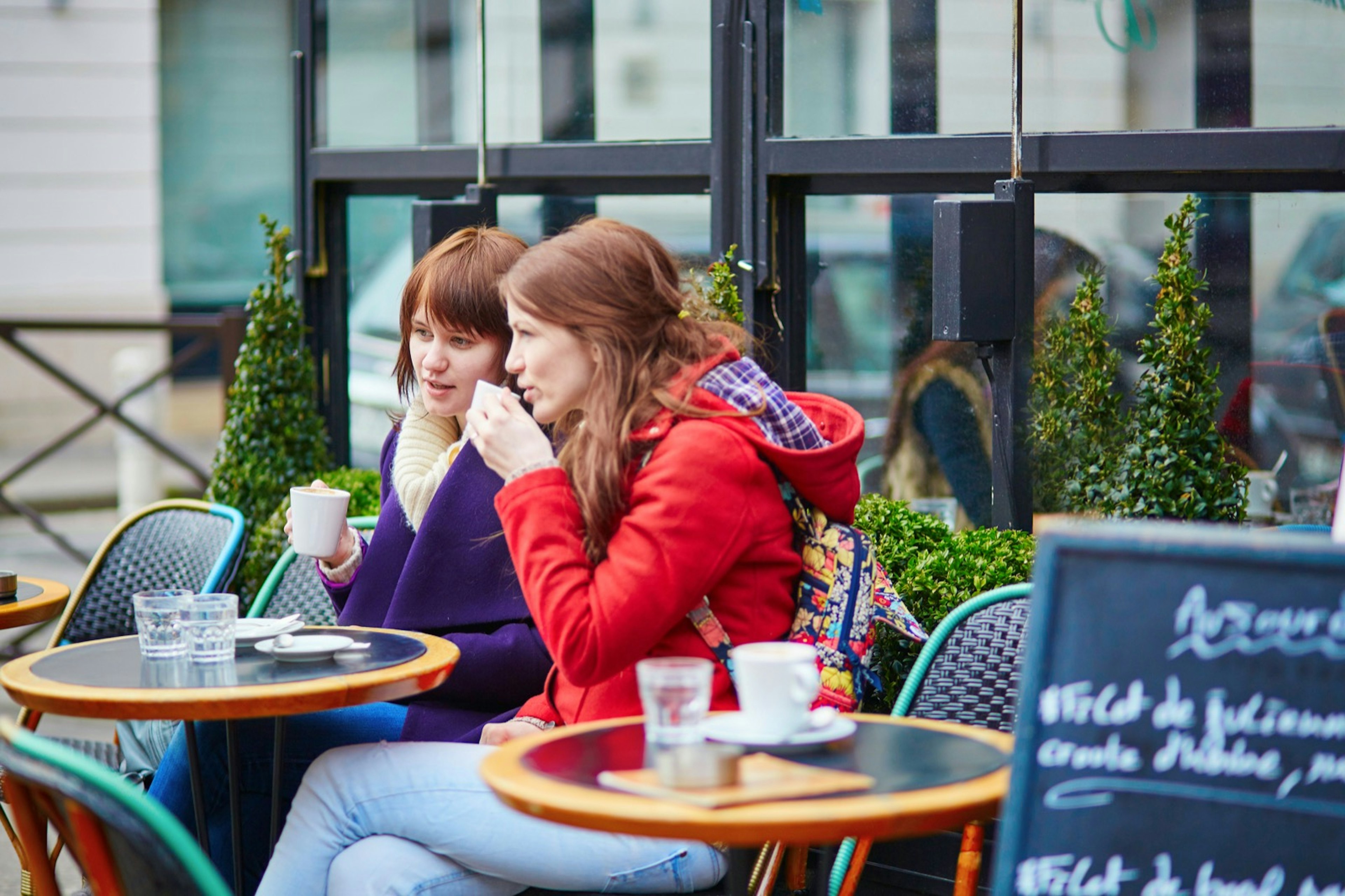 Two women sip drinks in coats at a street cafe in Paris