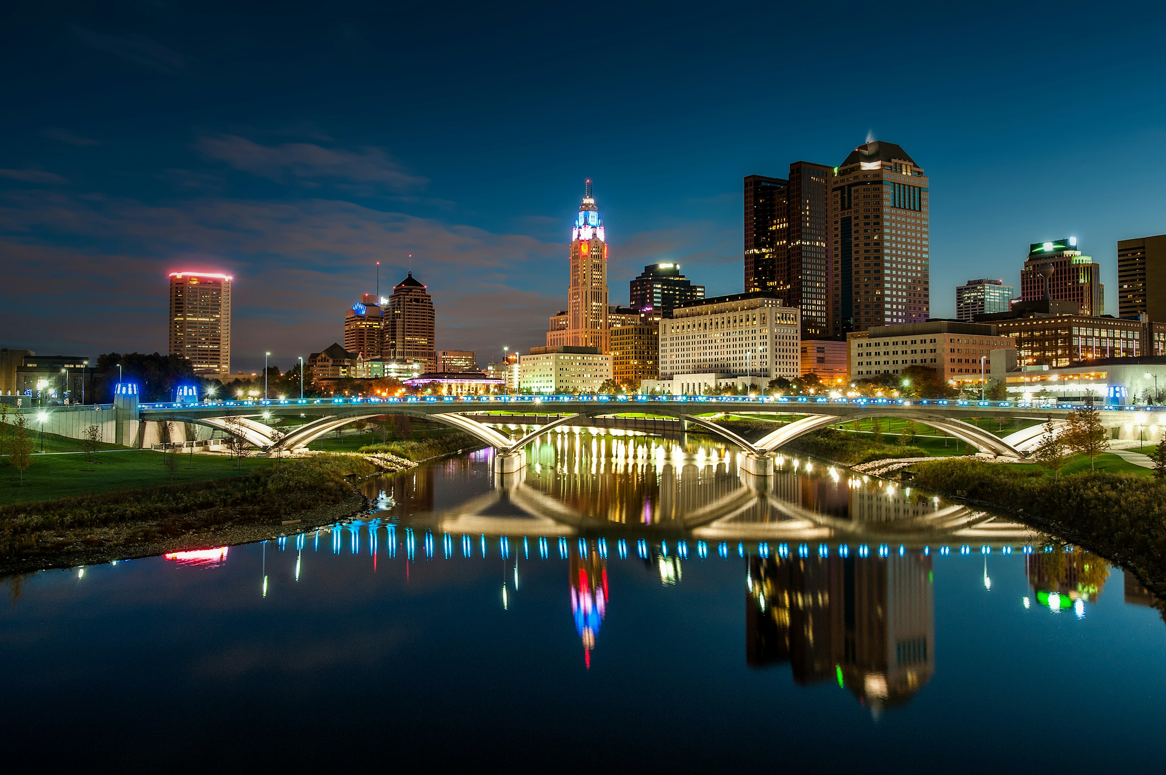 The bright city lights of downtown Columbus is mirrored in a body of water under flowing under a stone bridge; Columbus vs AnnArbor