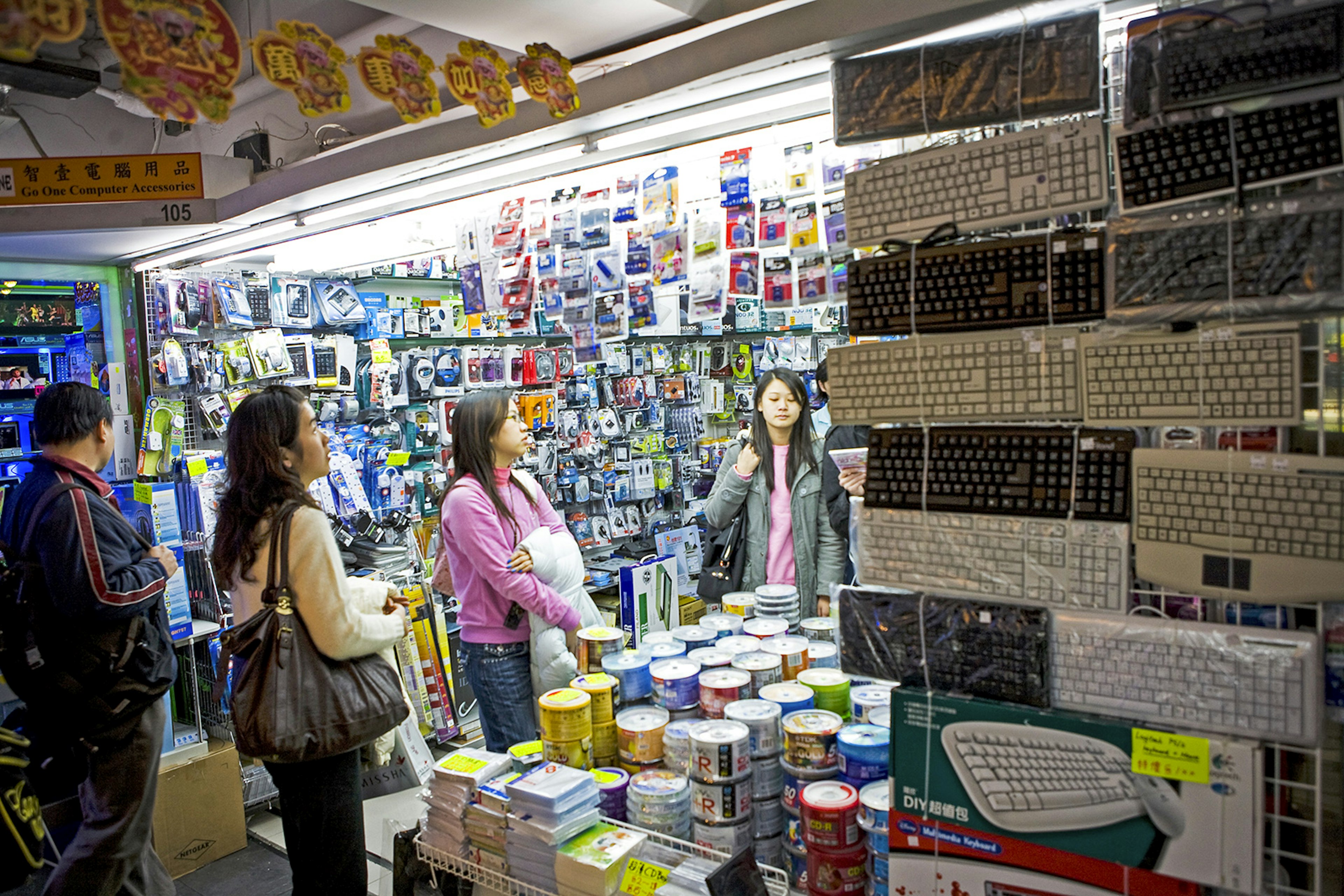 Three people stand in a well-lit stall with walls lined with electronics; keyboards are exhibited in the foreground. Hong Kong.