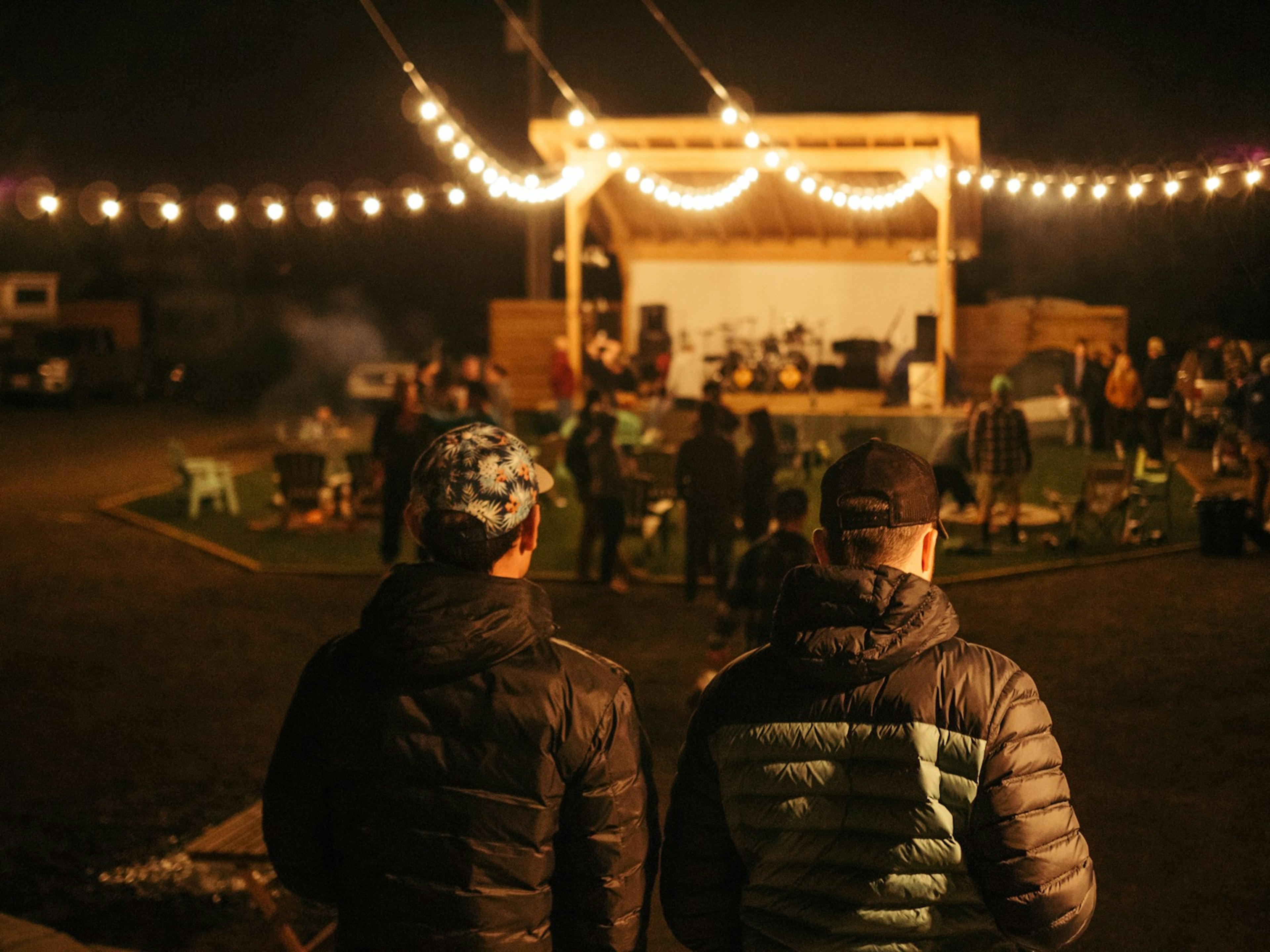 Two men in outdoors gear walk toward a small stage at night, as strings of globe lamps light their way