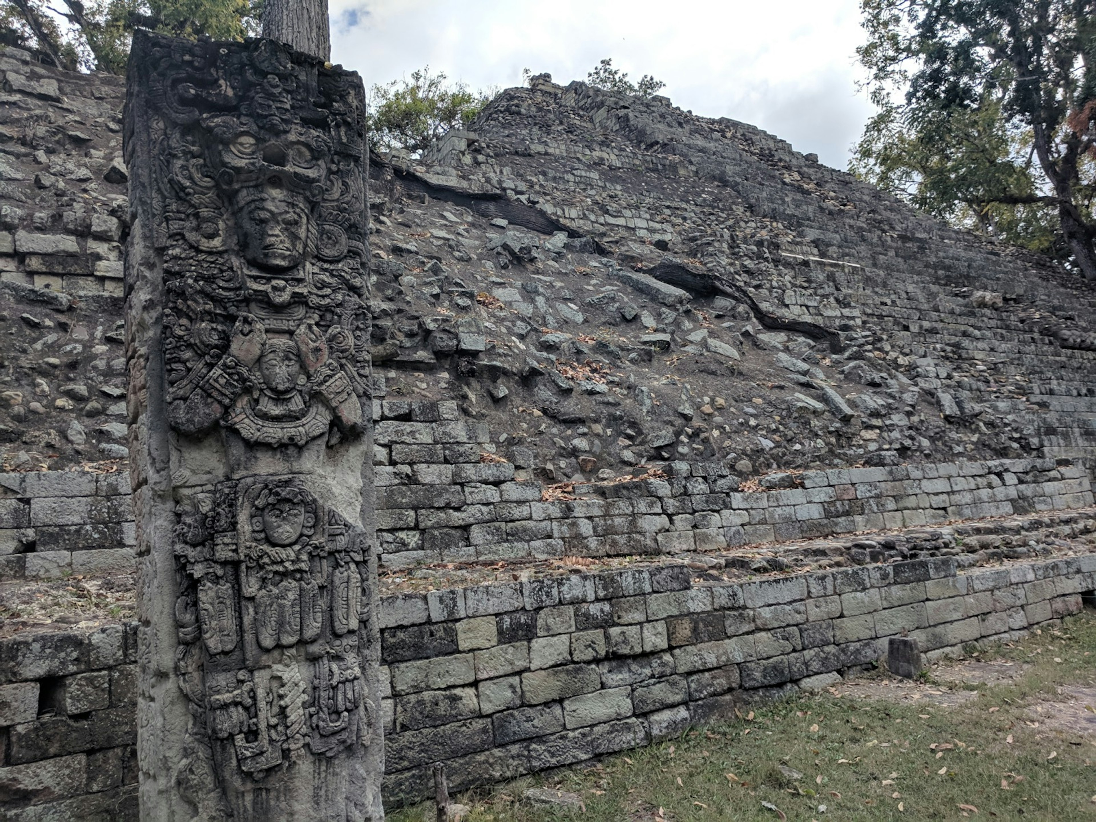 A mayan stone statue full of ornate carvings, thought aged and stained black by weathering, stands in front of crumbling stone pyramid at Copan Ruins in Honduras