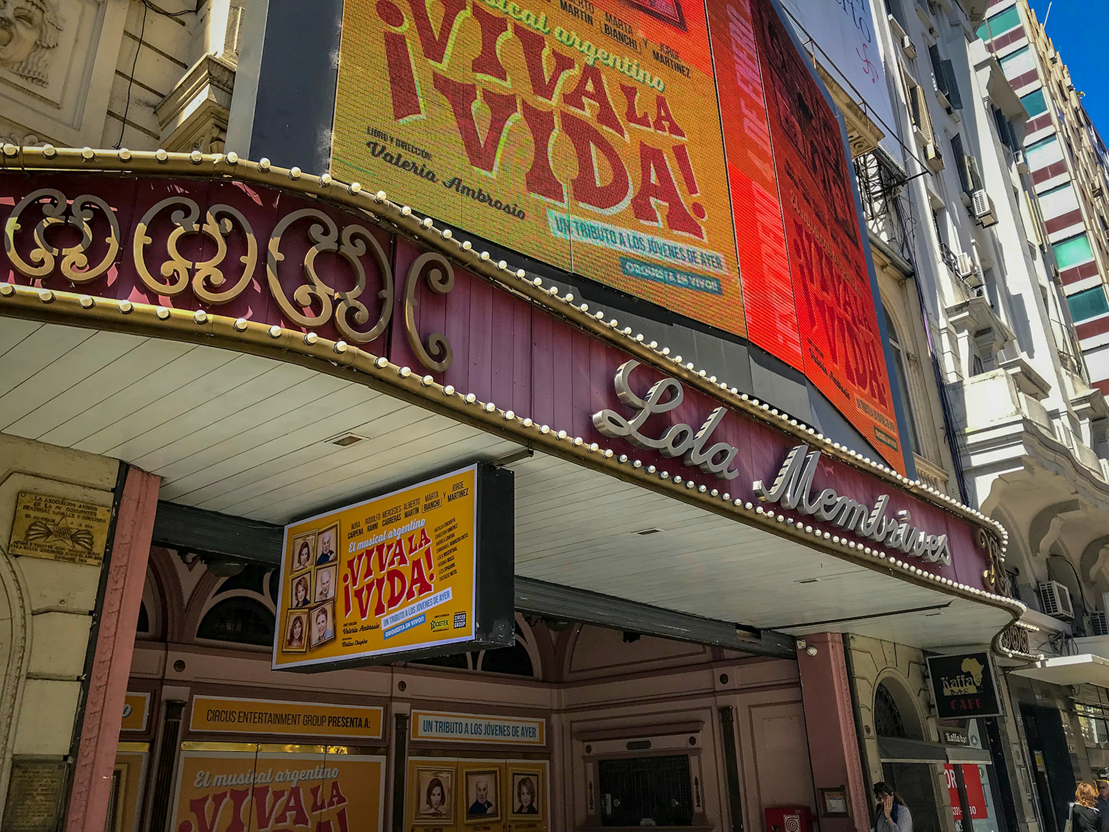 A purple marquee of a theater on Avenida Corrientes with its name written in cursive silver script; above is a billboard advertising a show titled