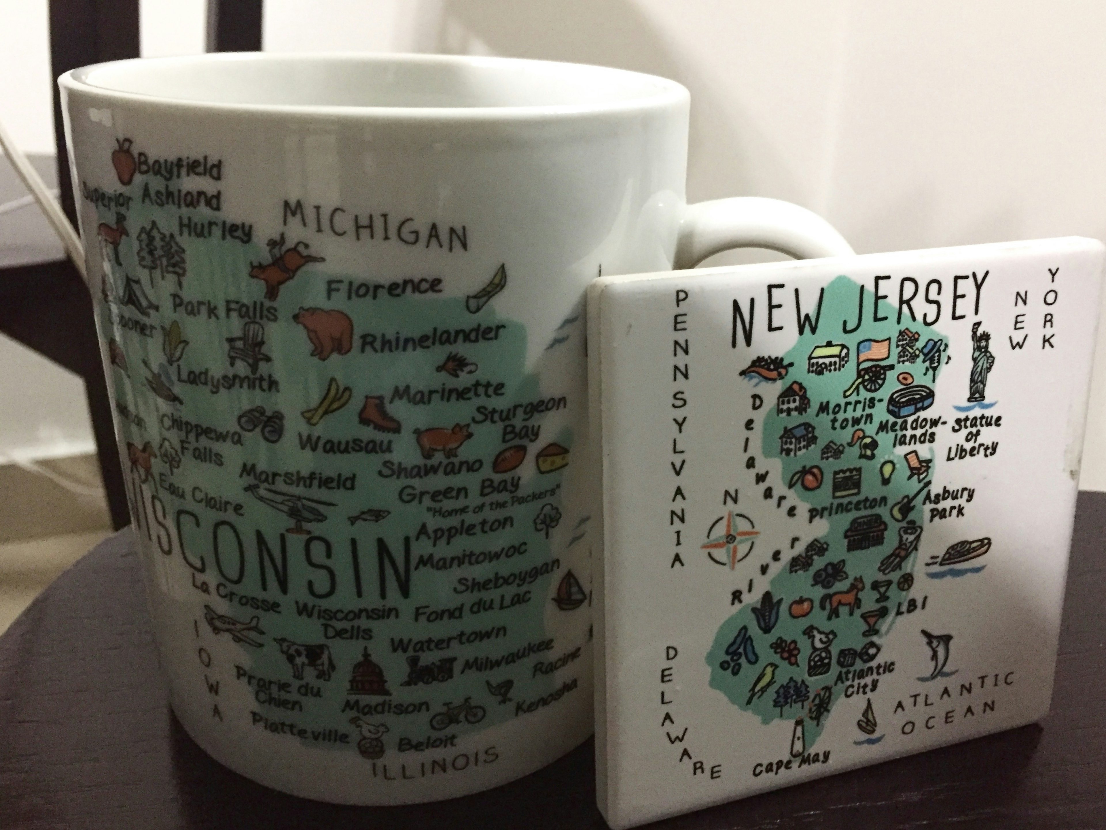 Closeup of a white tea cup etched with various cities and small images for the state of Wisconsin. Next to the cup is a white New Jersey coaster of the same design with various cities and images dotted throughout an outline of the state