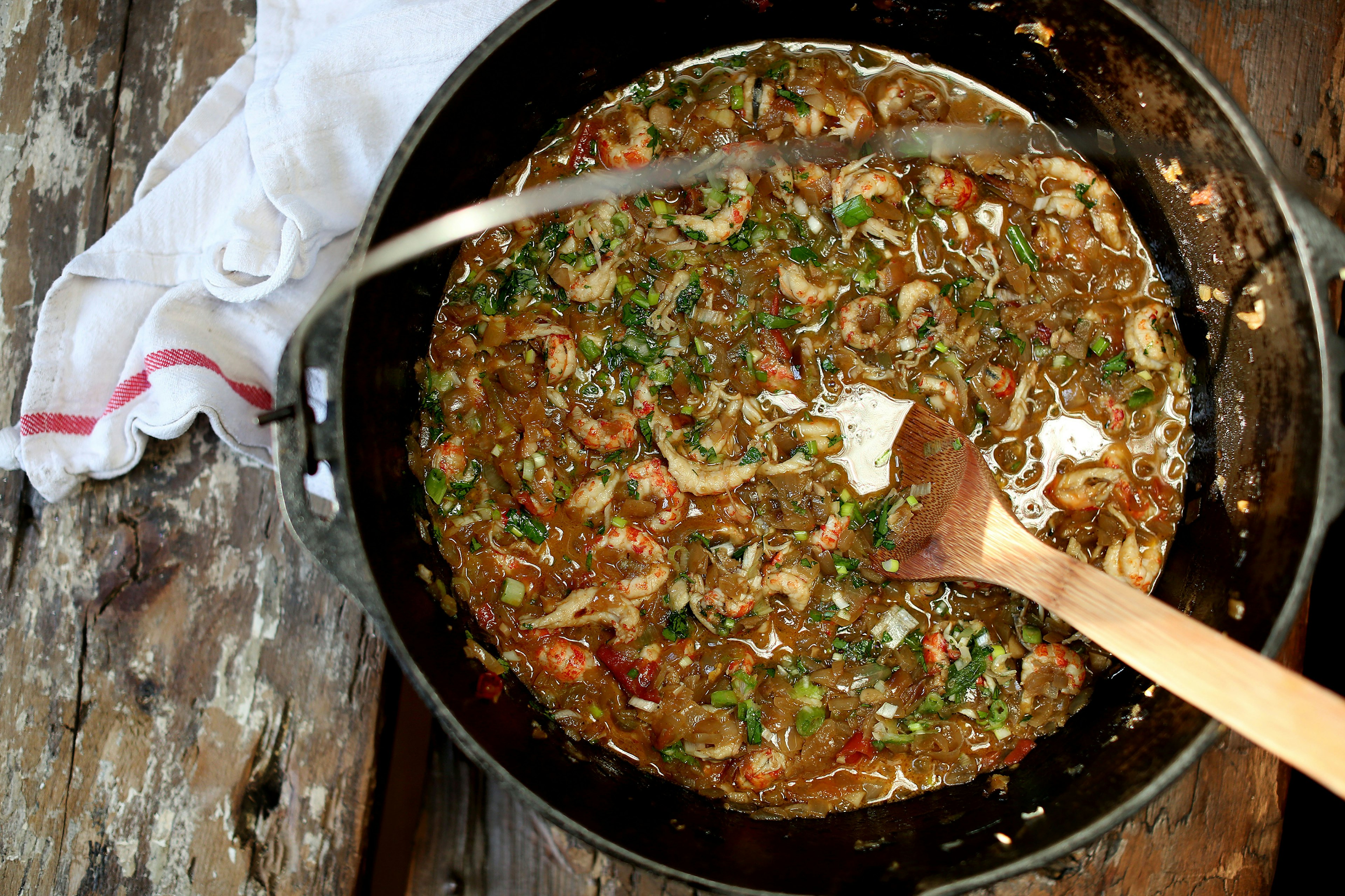 A wooden spoon sits in a large cast-iron pot of crawfish etouffee. There is a white towel with a red trim at the base of the pot.