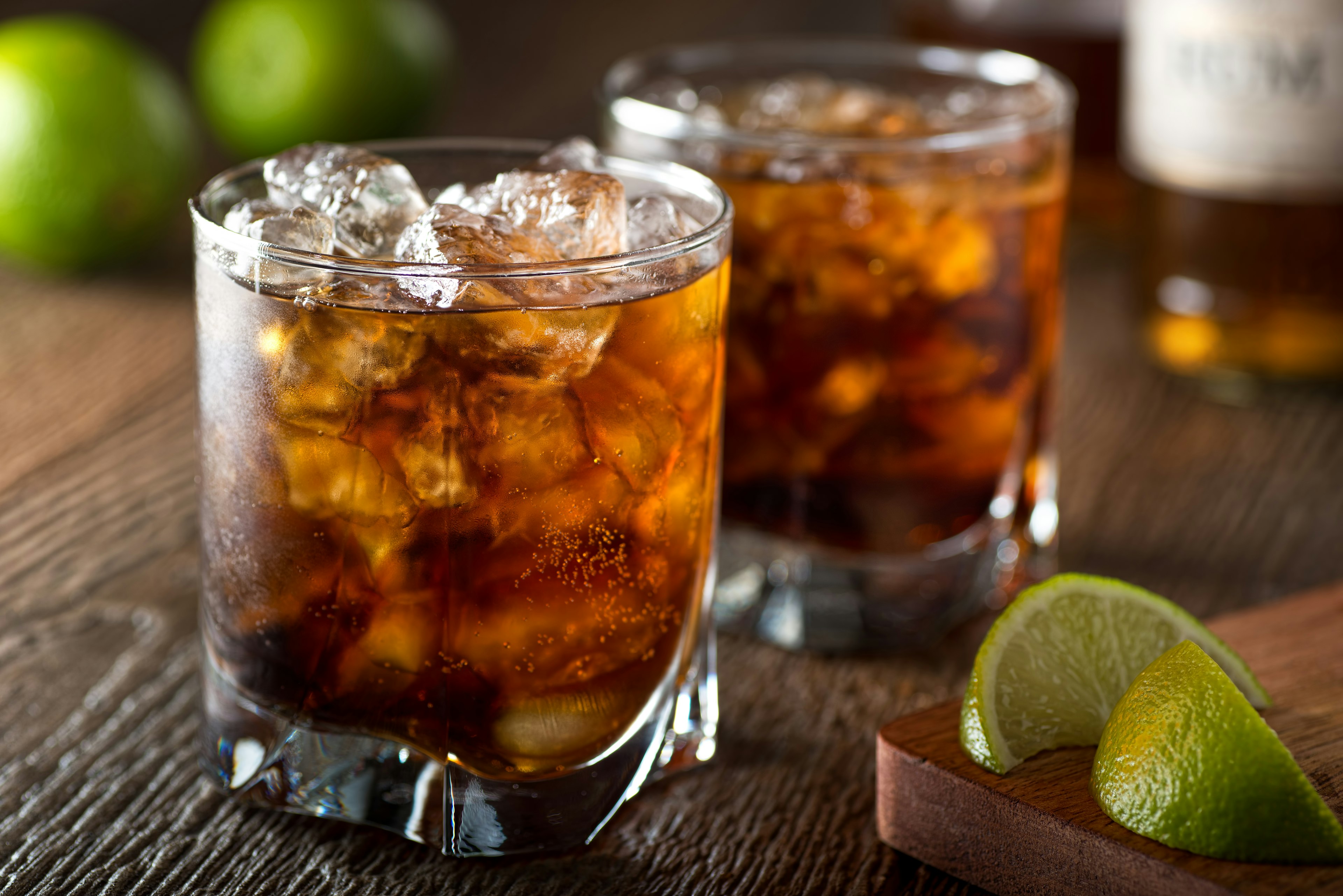 A pair of glasses filled with coca-cola, rum and ice to make a Cuba Libra, rest on a wooden table. On the left are a pair of lime wedges laying on a wooden cutting board. In the background is a bottle of rum and a pair of limes.