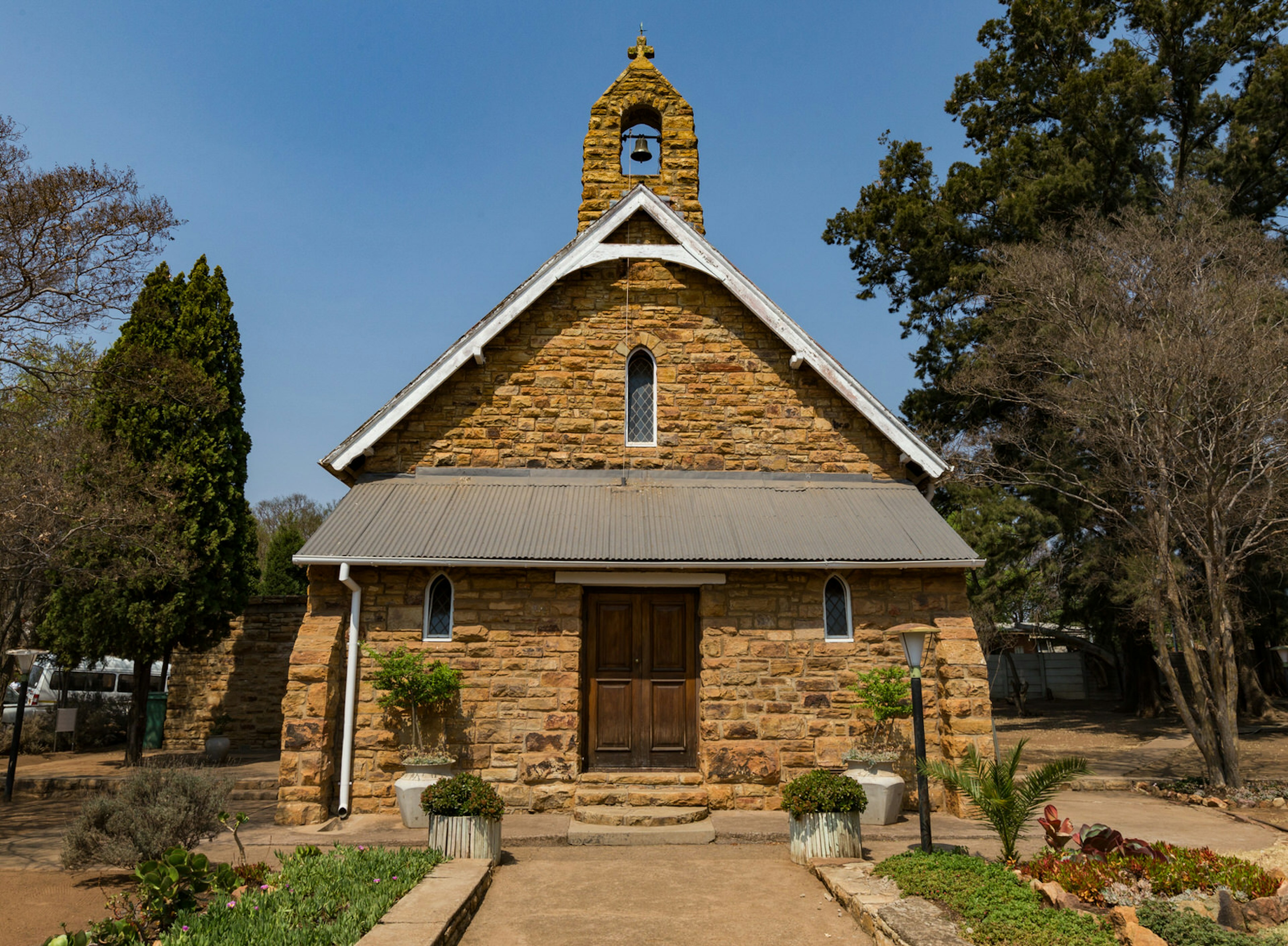 Under a blue sky is a A-framed rock church of yellow-brown hues; atop the church is a rounded tower with the middle cut out to hold a bell