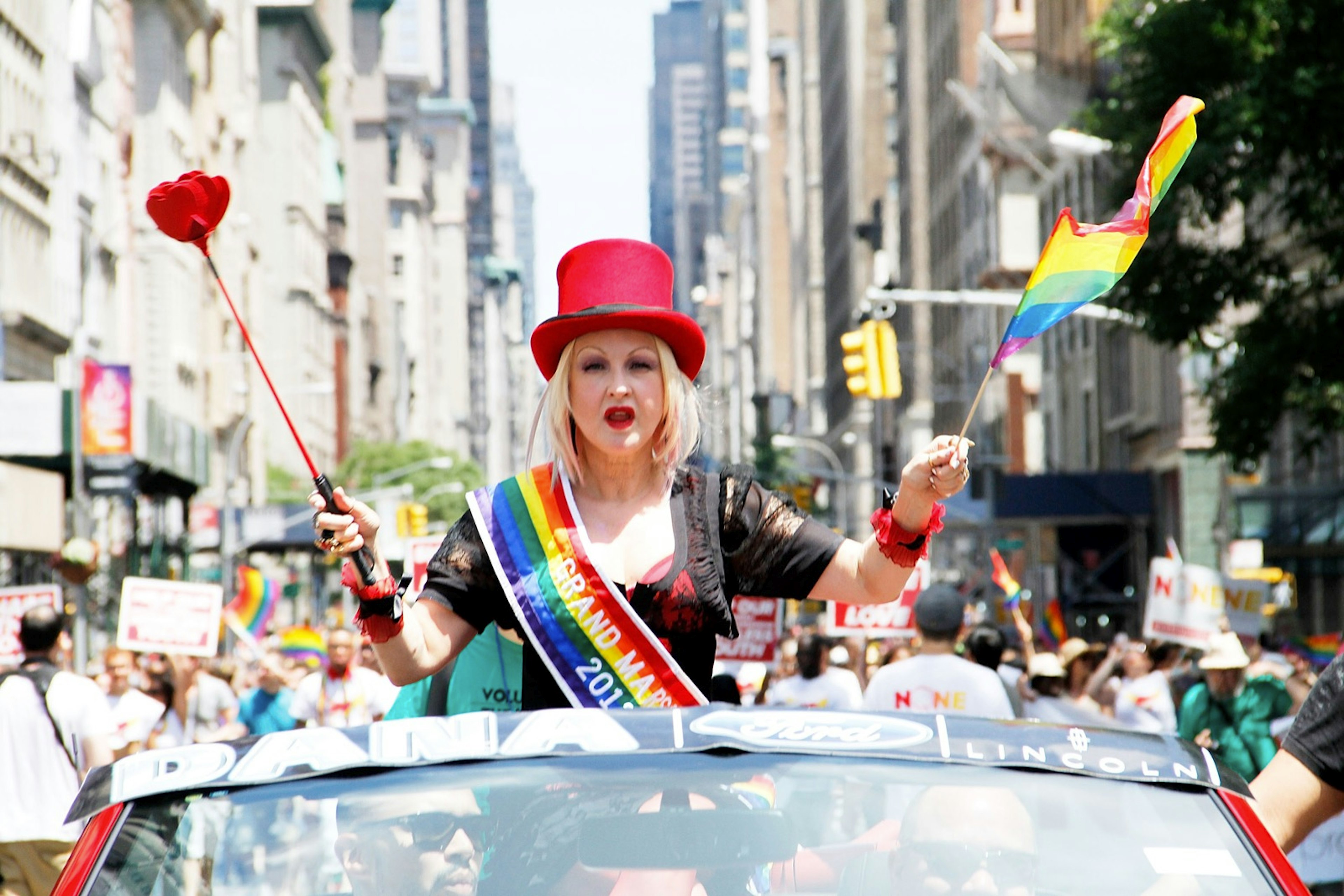 Musical artist Cyndi Lauper wears a red top hat while holding a rainbow flag and a large plastic rose as she sits in a convertible car during the New York City Pride parade. Lauper will be among the many musical acts performing at NYC WorldPride.