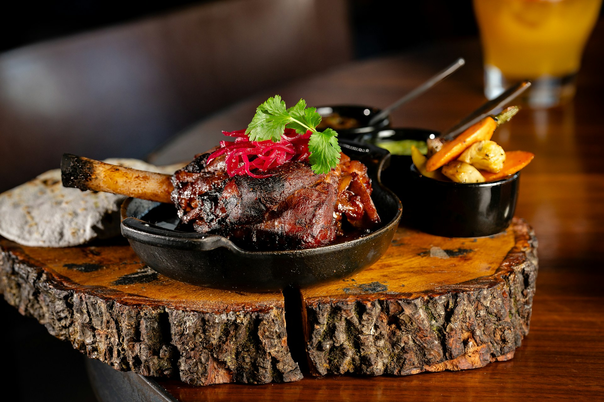 A lamb shank sits in a cast iron skillet topped with vegetables and cilantro. Vegetables in small side dishes sit behind. 