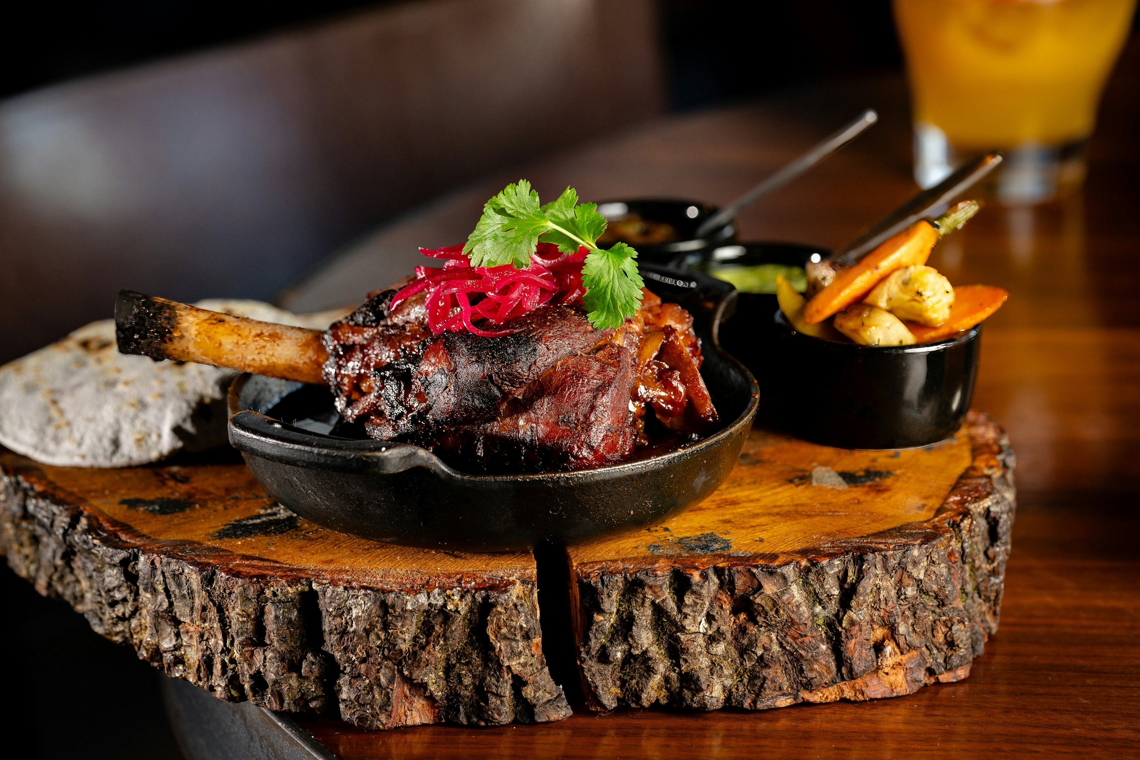 A lamb shank sits in a cast iron skillet topped with vegetables and cilantro.