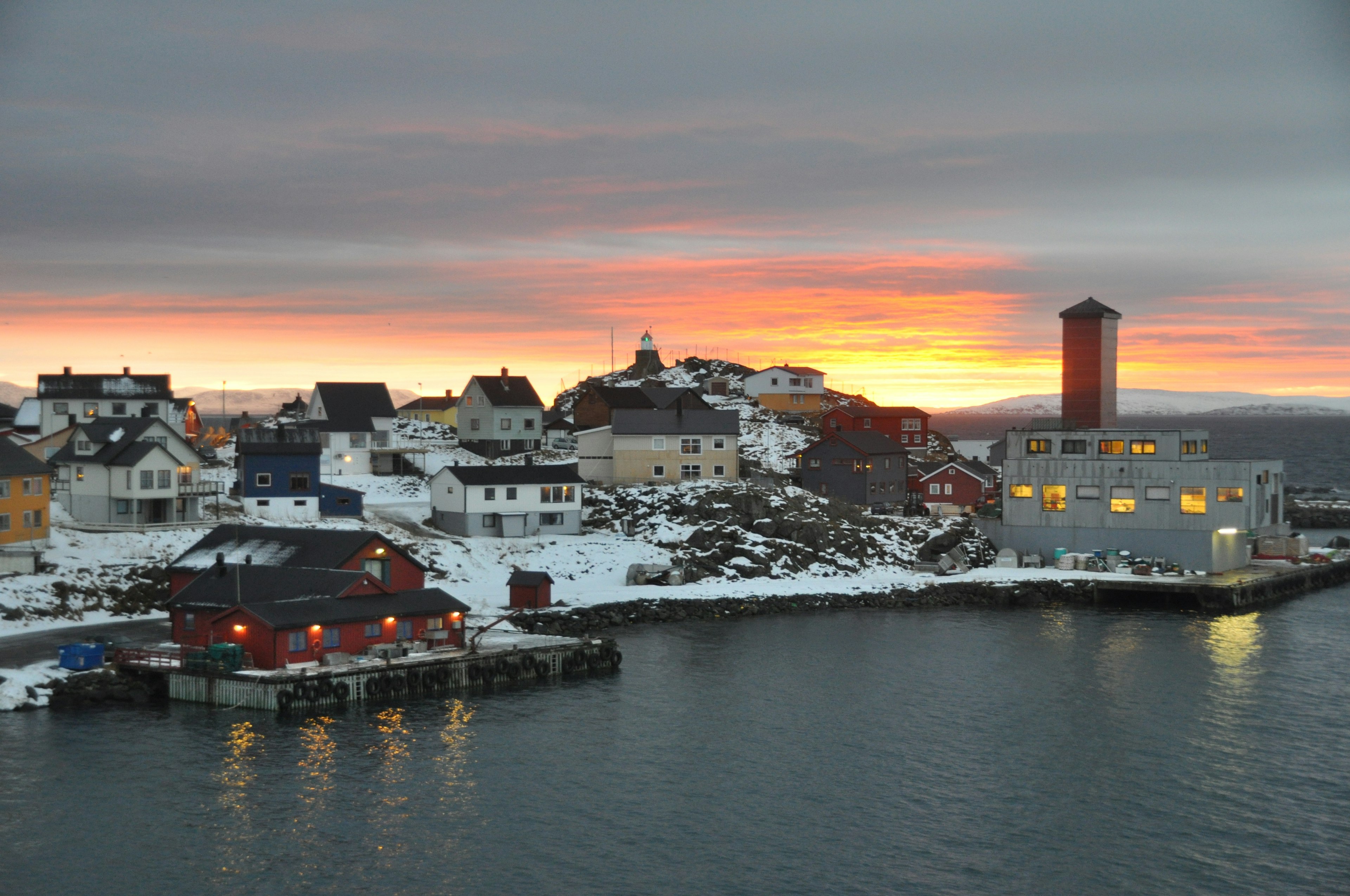 The port of a small coastal town in Norway with an orange sunset and buildings dusted in snow.