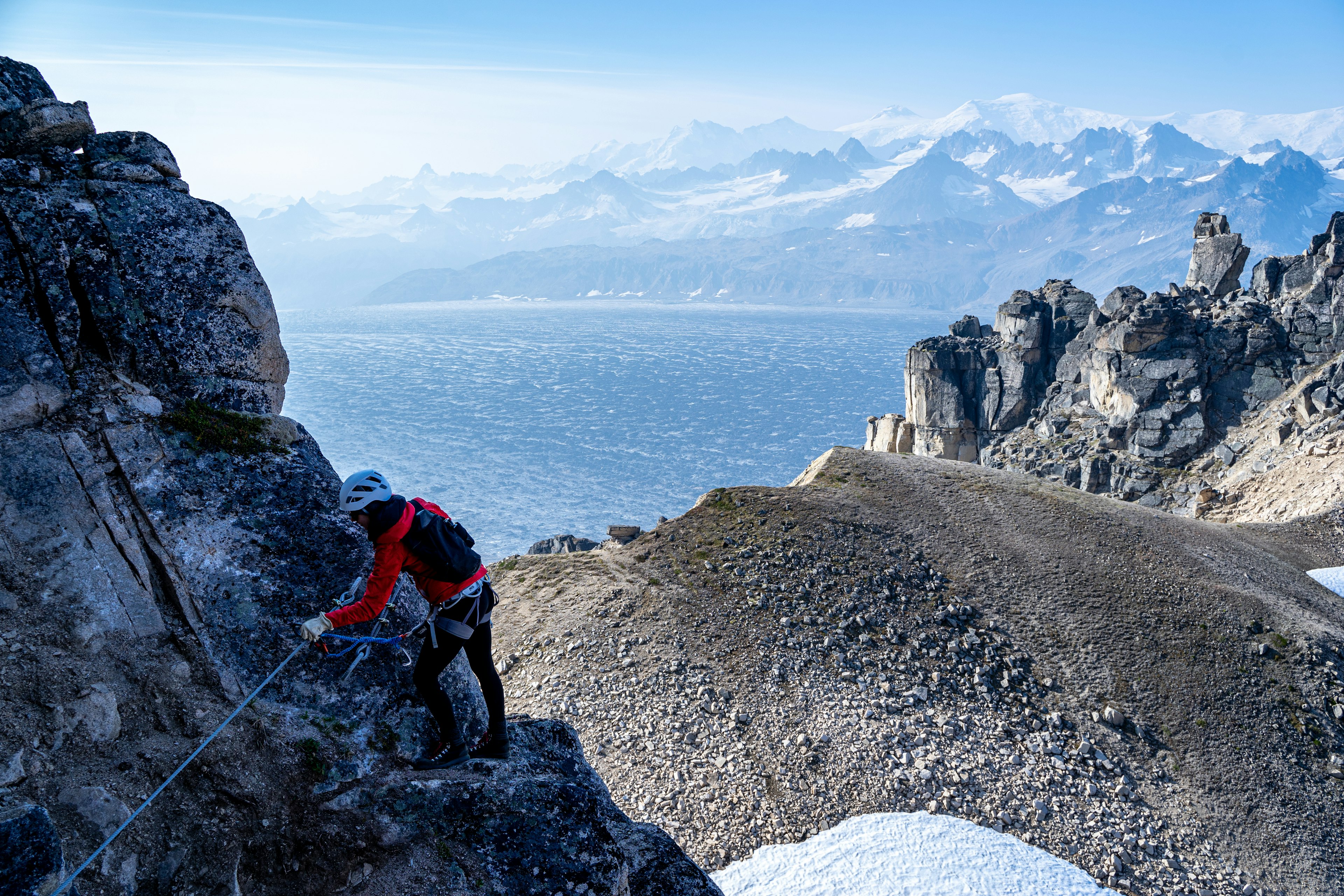 A woman picks her way along a rock wall with a mountain view and ocean in the background