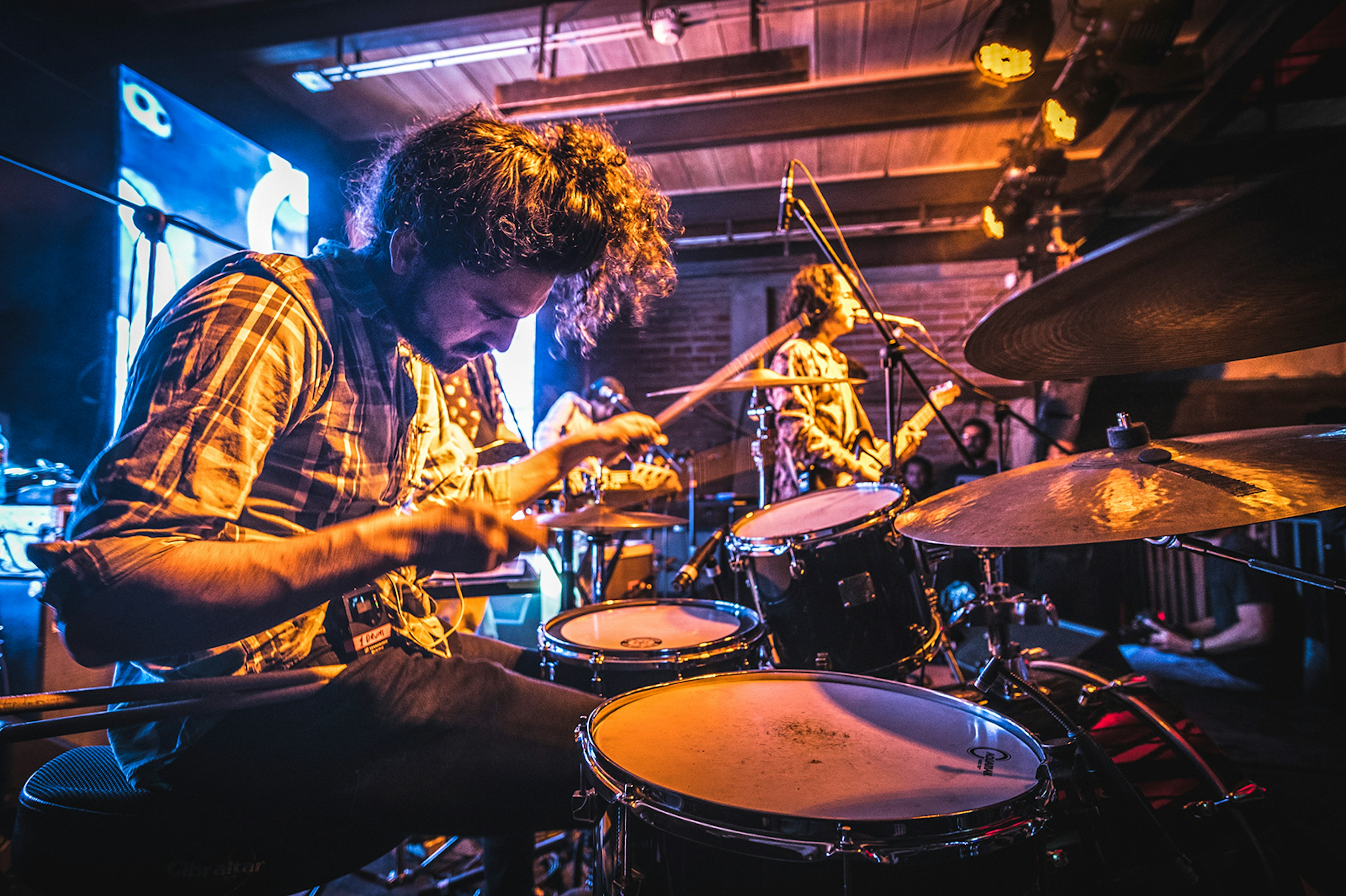 Close-up of drummer playing in a dark music venue with singer in the background © Da Pawn