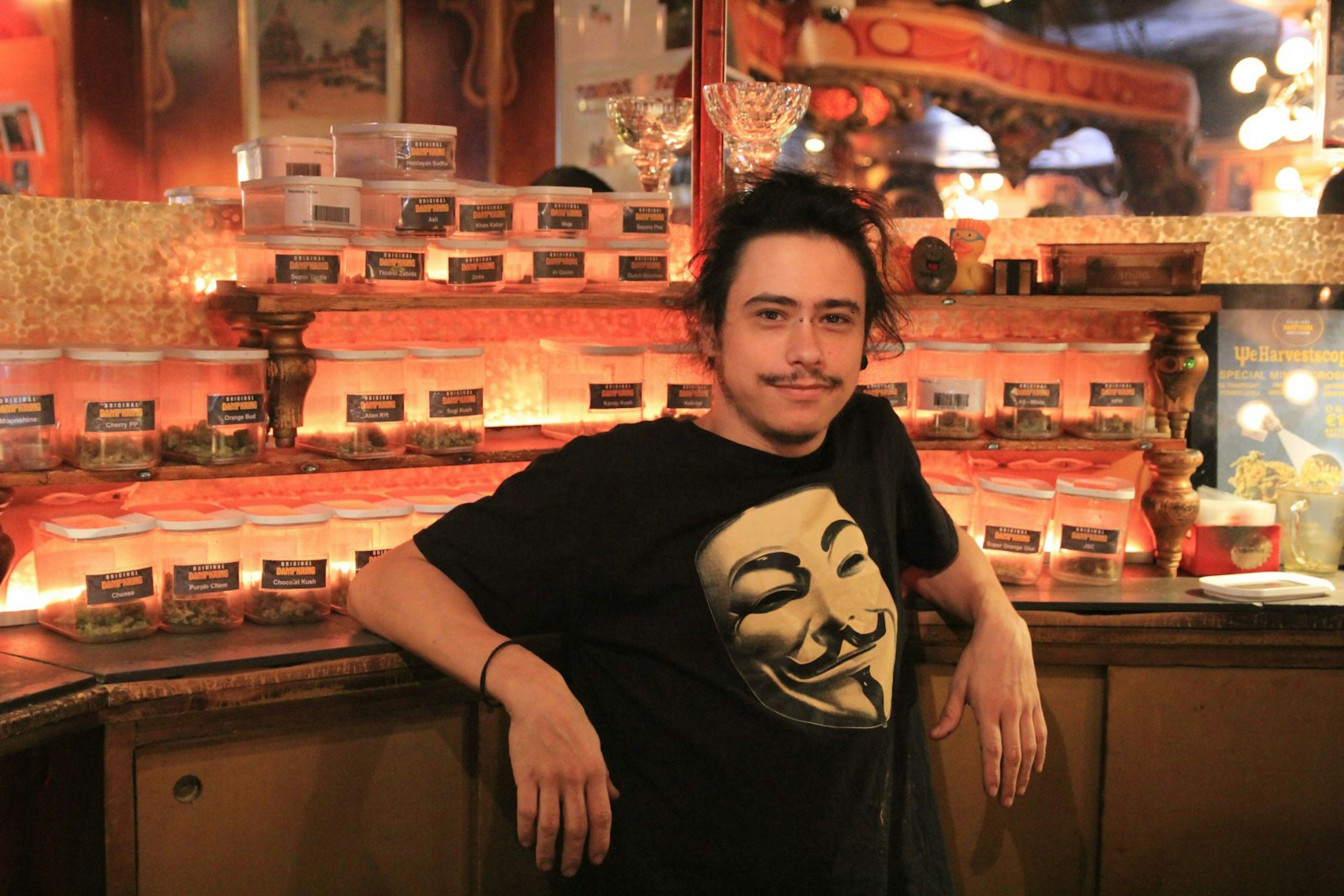 A man leans against the counter in Dampkring coffeeshop, Amsterdam. He is wearing a t-shirt with a mask on it and smiling at the camera. Behind the counter there are lots of containers with different kinds of weed.