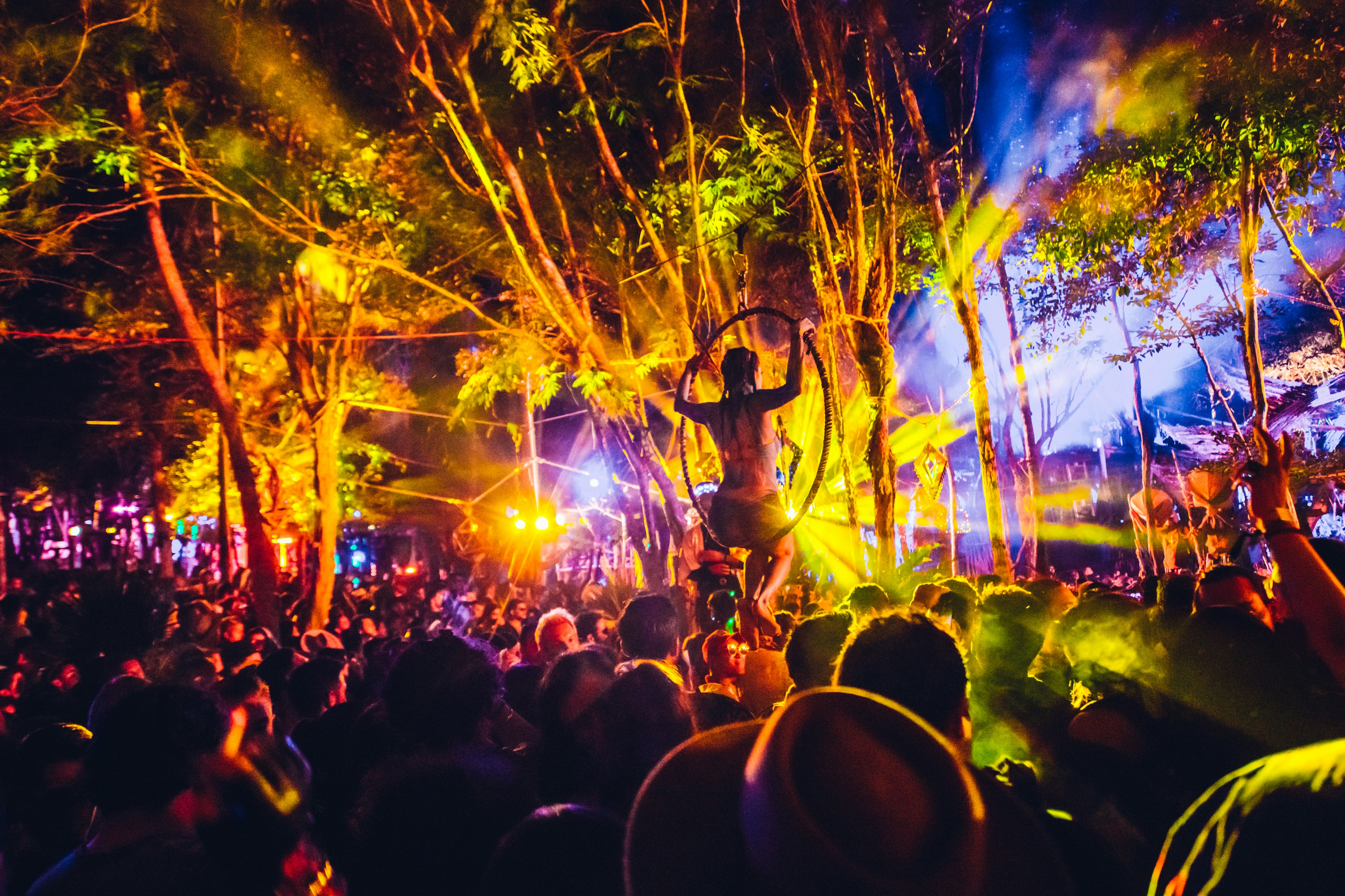 a huge crowd is gathered in front of a stage at night time. Multi-coloured lights are beaming from the stage illuminating the crowd including a performer suspended over the crowd in a hoop at Day Zero Festival in Masada, Israel.