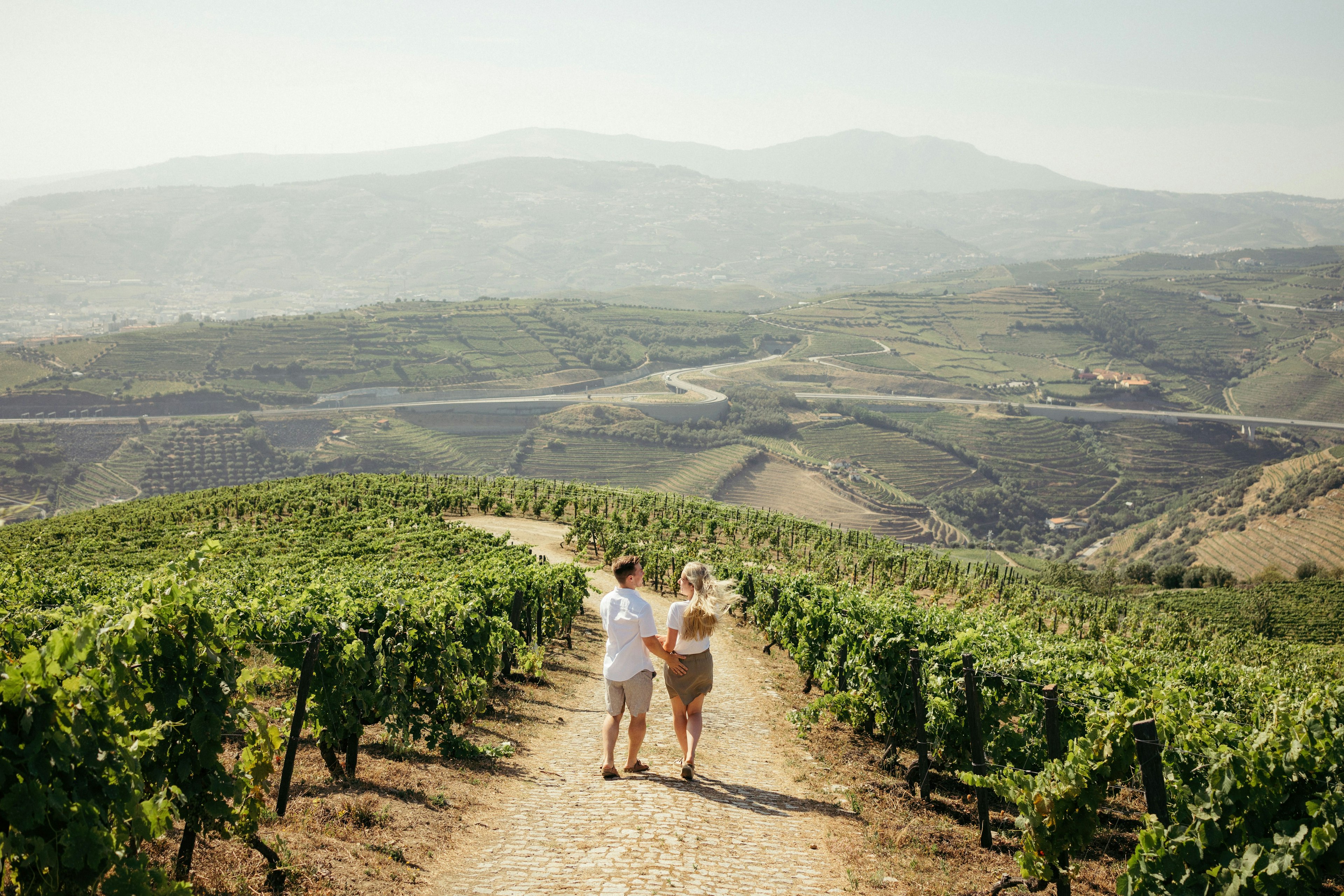 A couple walk arm-in-arm down a hill in the countryside with sunlit fields stretching out in front of them.