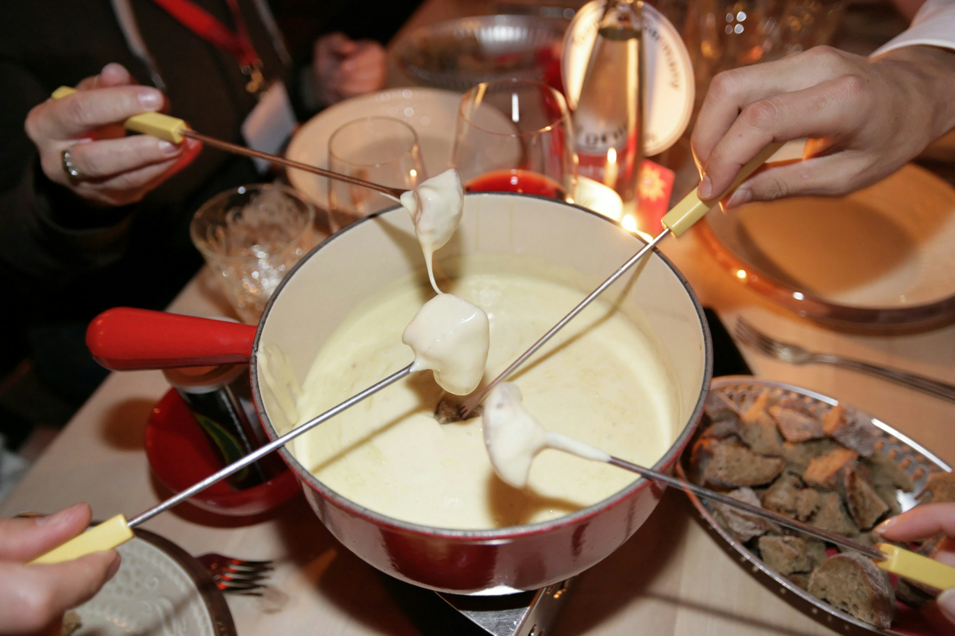 Several diners are dipping pieces of bread into a delicious cheese fondue with wood and metal fondu forks. The cheese is white and melty and the fondu pot is surrounded by smaller dishes and glasses of red wine