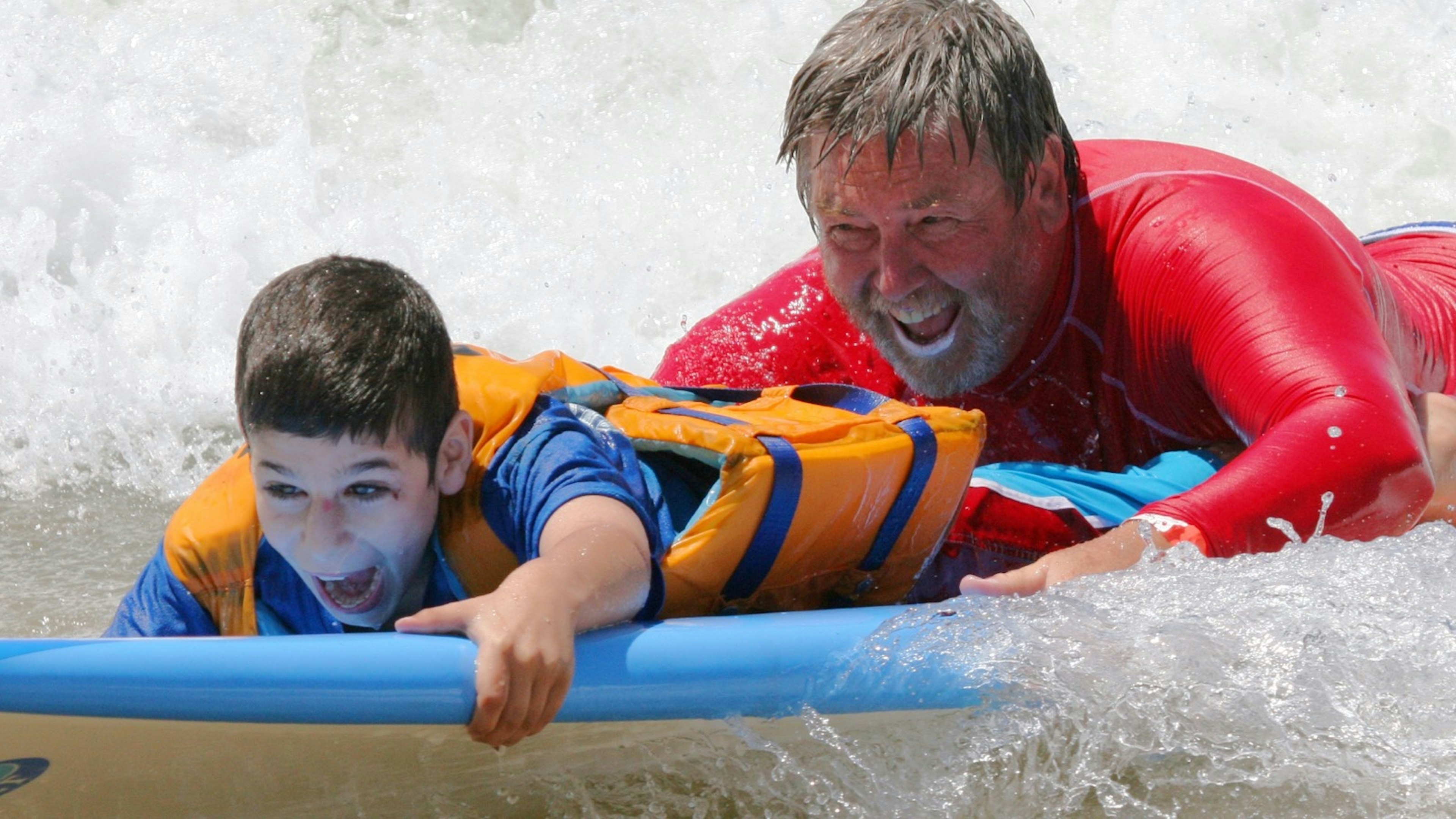 A boy in an orange life jacket smiles as a man in a red rashguard guides a surf board through the waves; accessible Australia