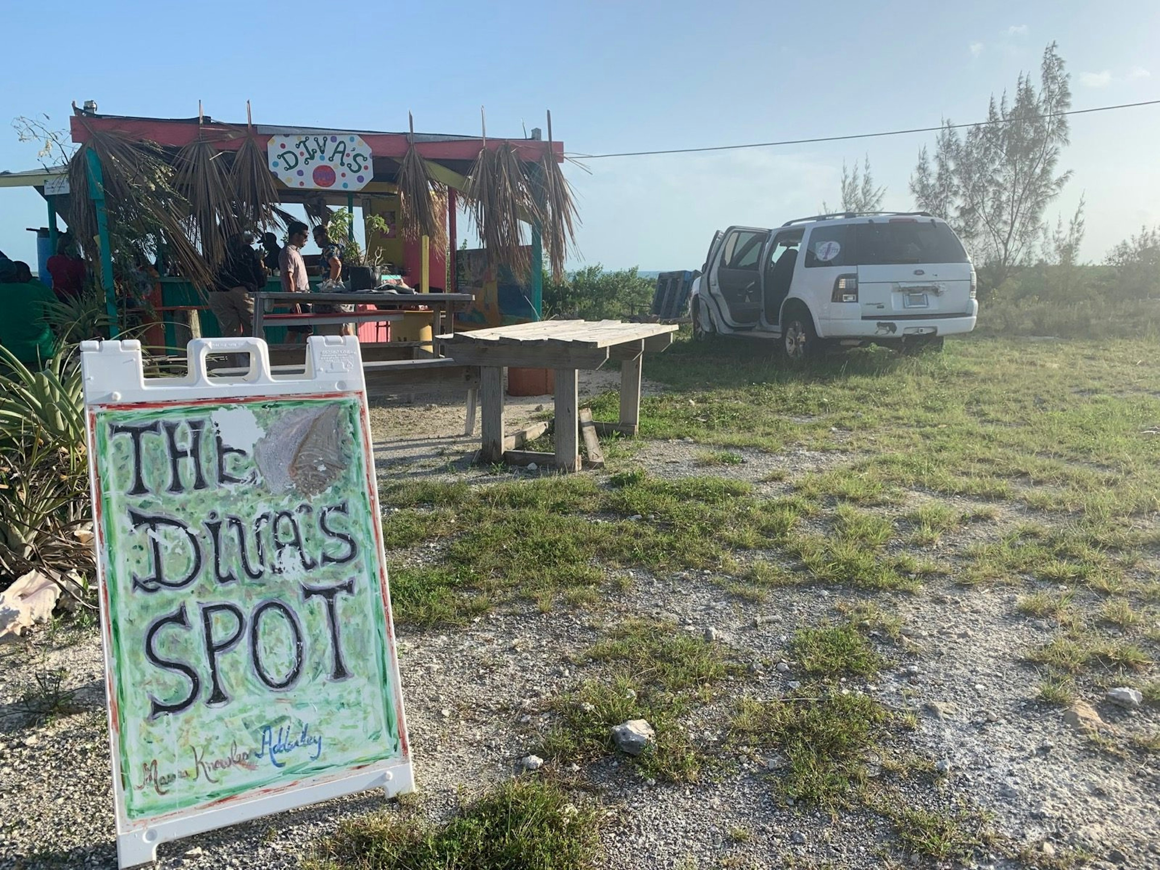 A sign with 'The Diva's Spot' sits in front of a table and wooden bar covered with palm leaves. There's a white SUV parked on the grass on the right; Long Island Bahamas