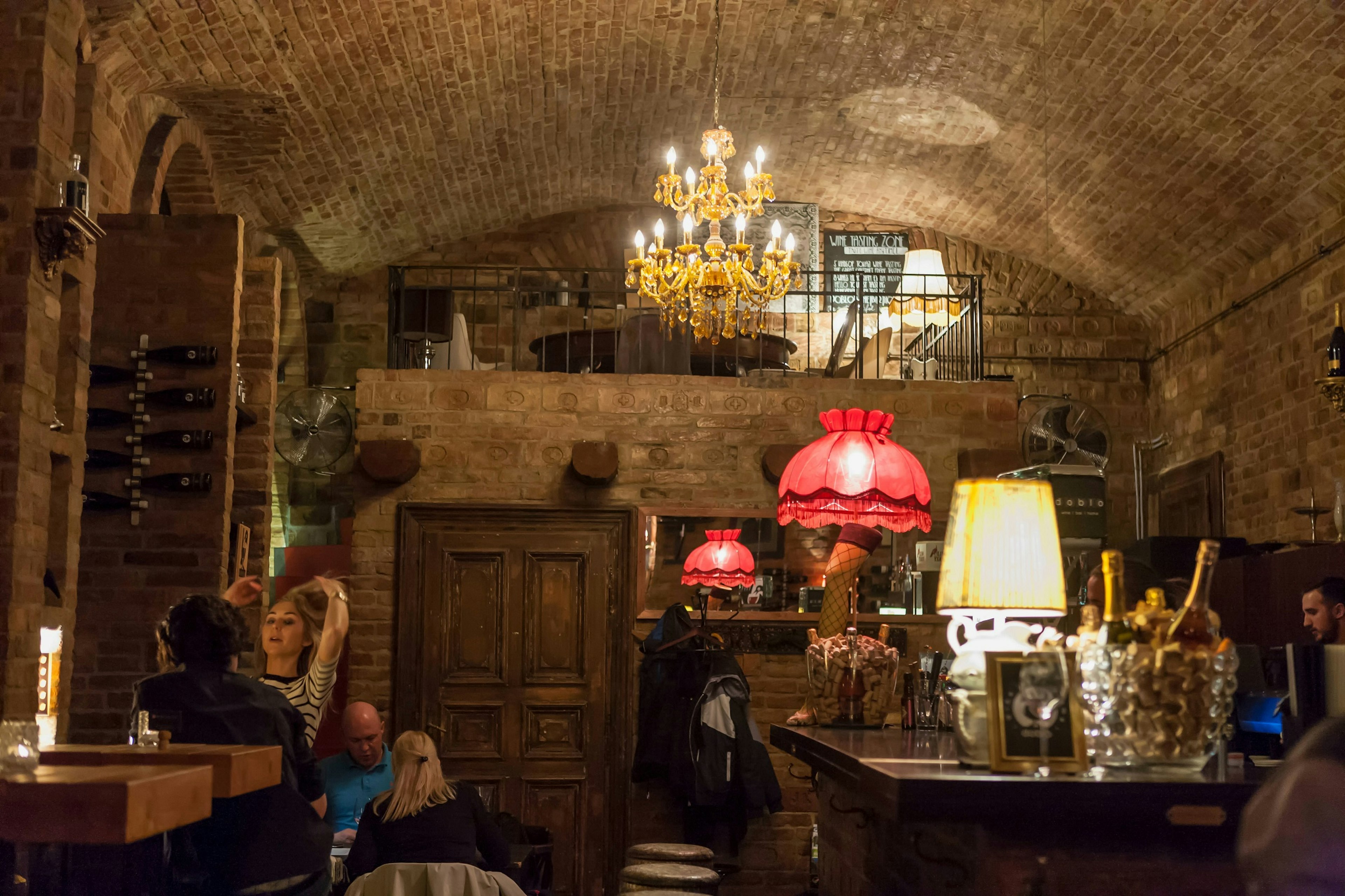 A cosy, cellar-like room with brick ceiling is lit by a chandelier and two standing lamps. Wine bottles can be seen on the bar to the right.