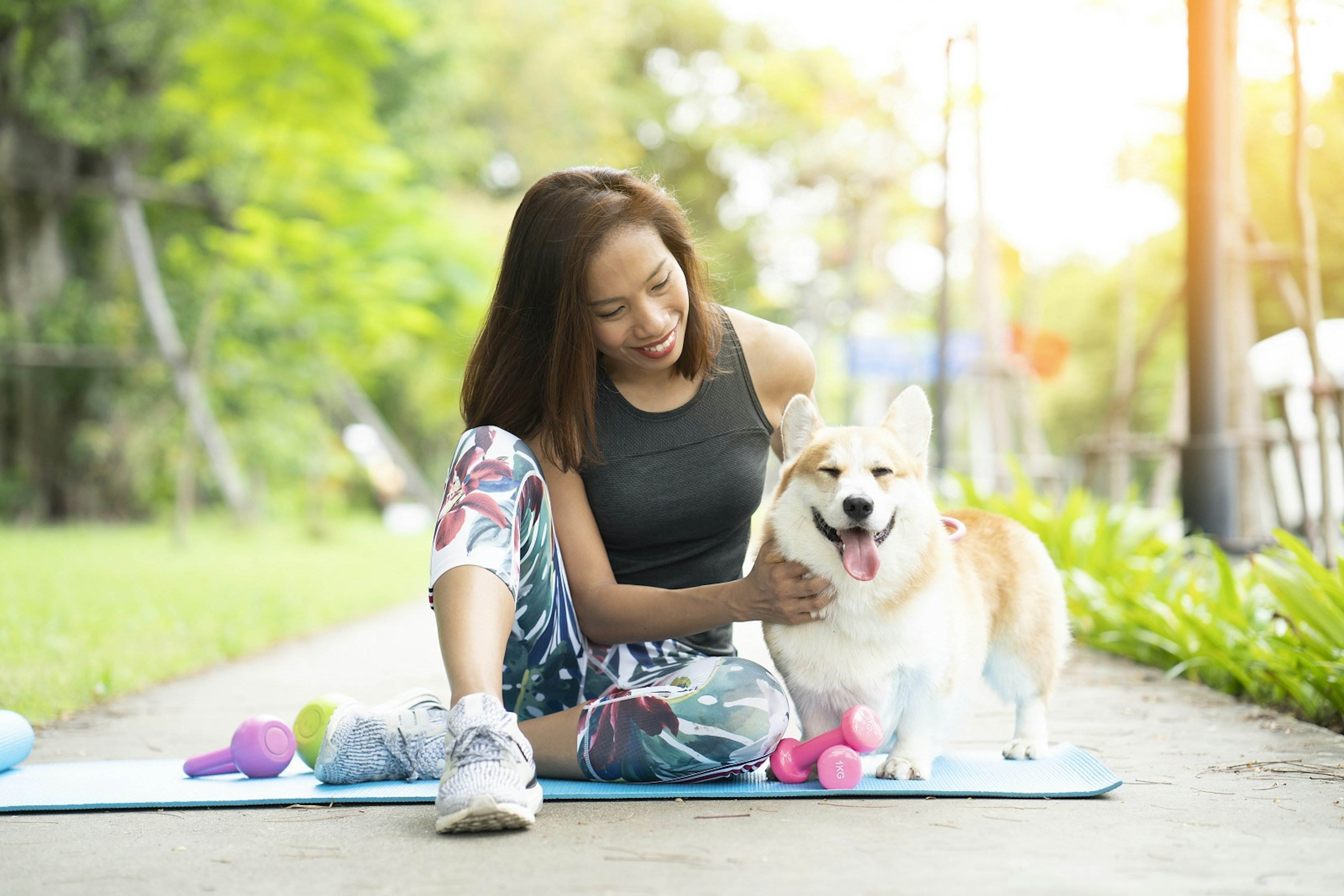 A healthy woman playing with a corgi puppy while exercising on a yoga mat surrounded with gym tools such as kettlebell and dumbbell in outdoor training.