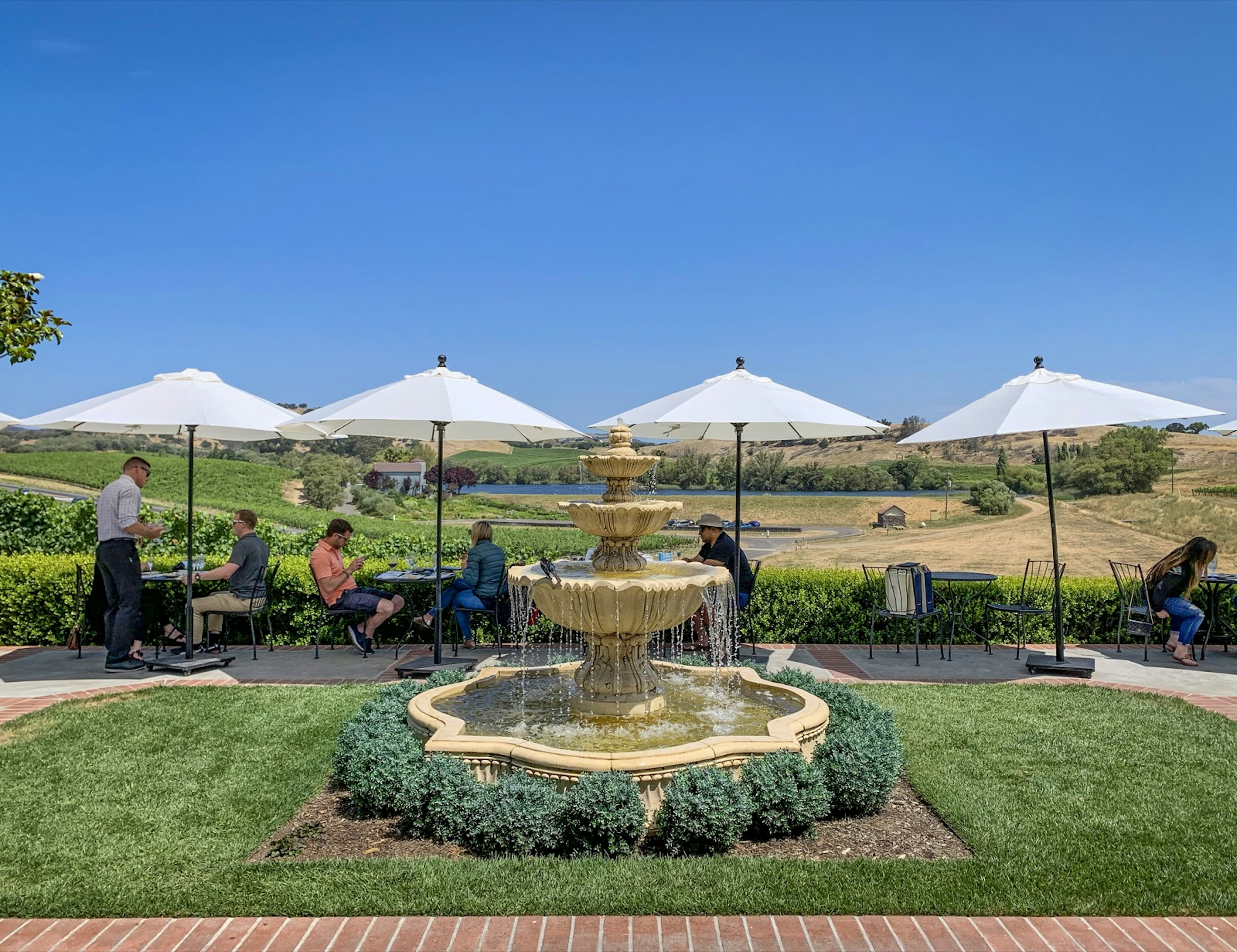 A collection of people sit at tables under umbrellas near a fountain; weekend in Napap