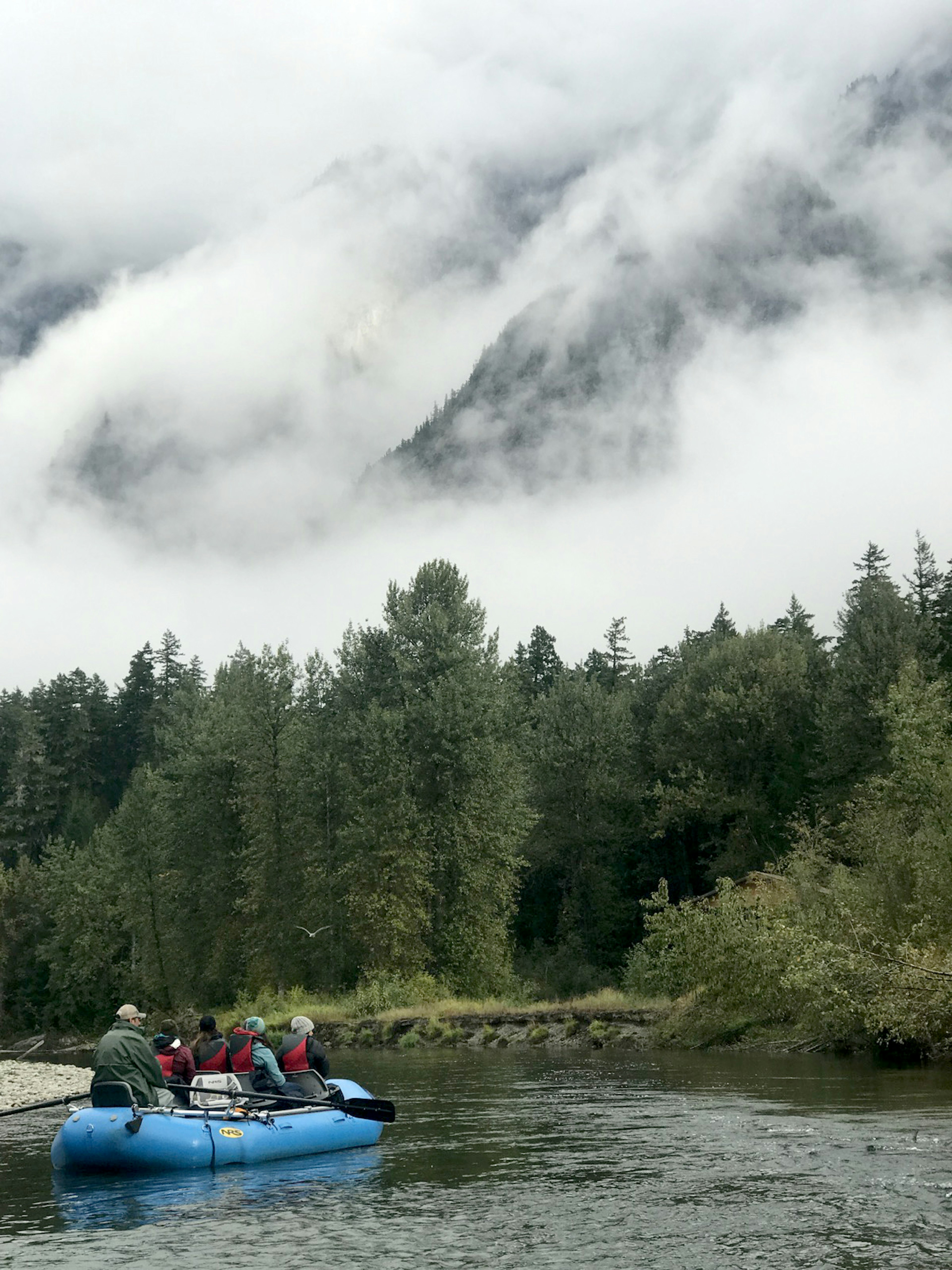 Clouds cling to the ridges of a mountain as an inflatable drift boat glides down a river in British Columbia.