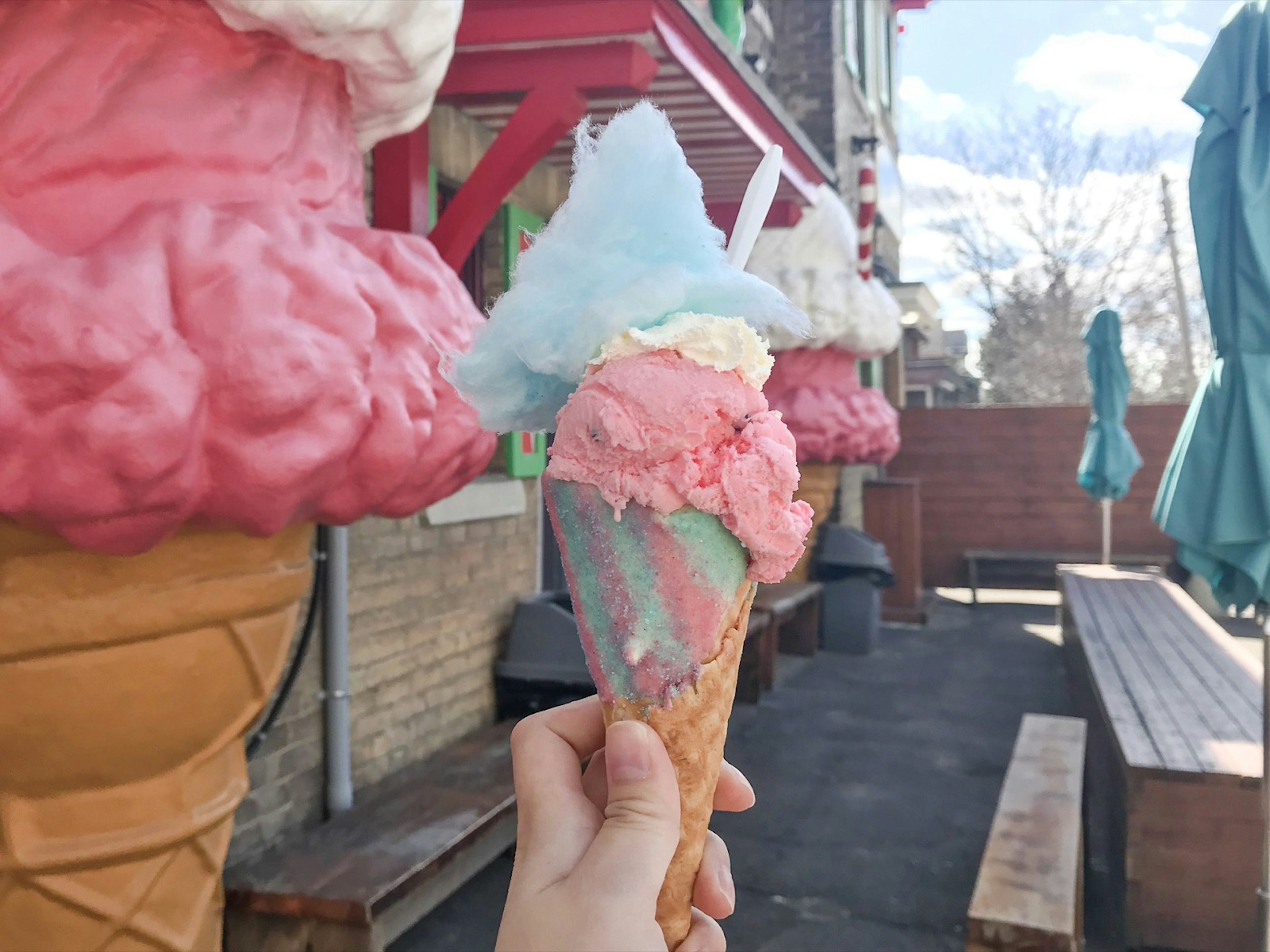 A pink and blue fancy ice cream cone is held in front of giant fiberclass cones at a shop