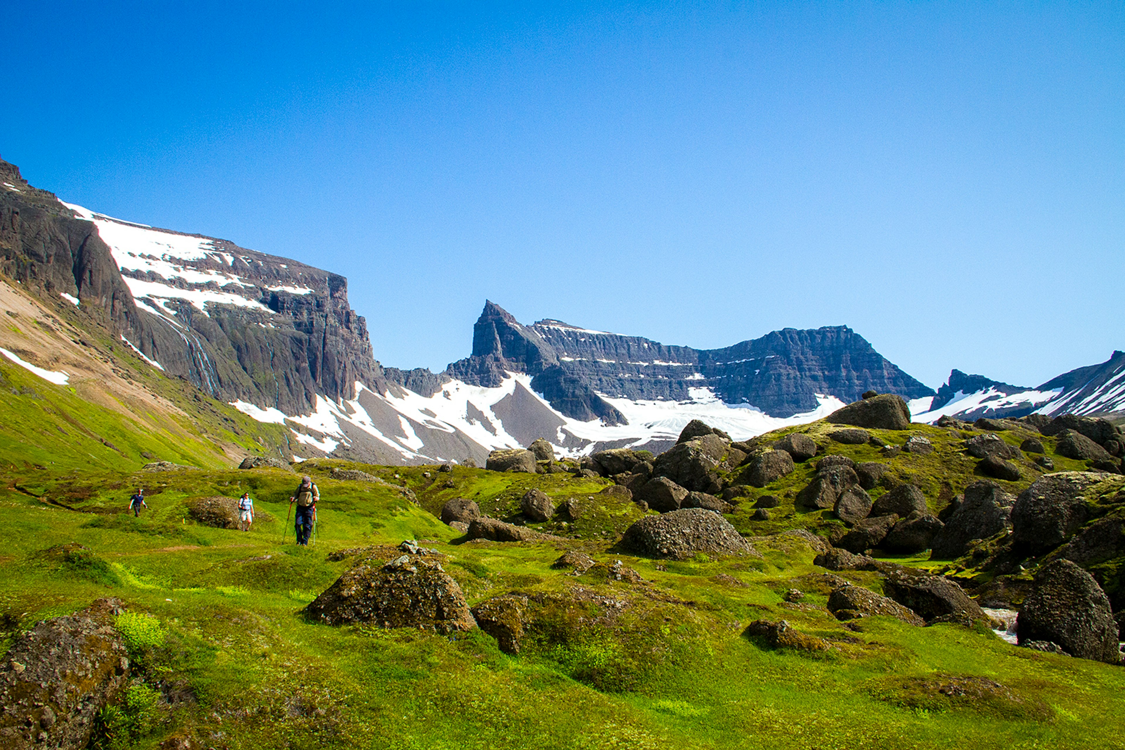 Hikers walk through mossy fields with a snowy rock ridge in the background; the ridge has an opening in the middle. East Iceland.