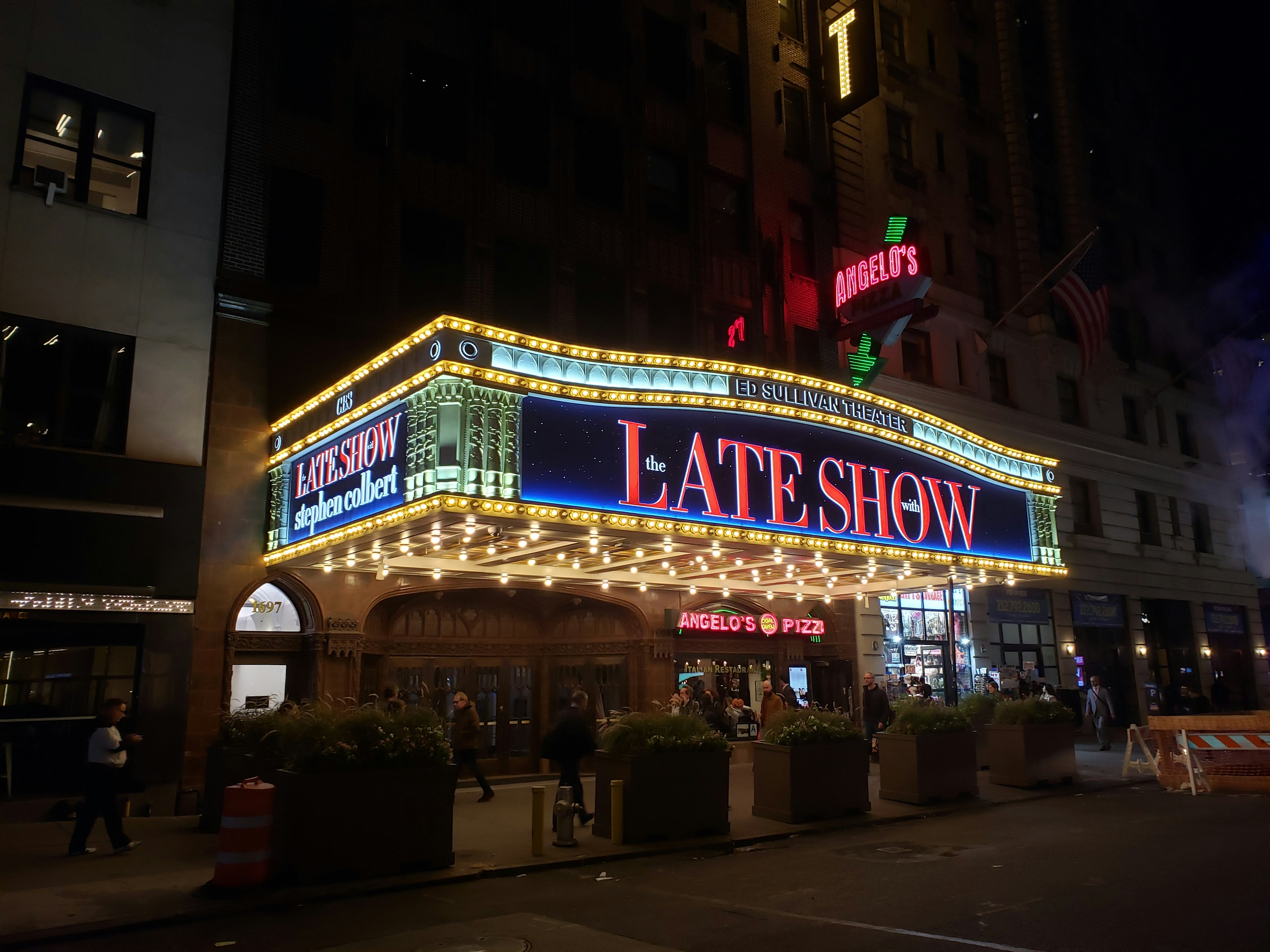 The exterior of the Ed Sullivan Theater shows a vast overhanging marquee with large-bulb theater lights on the underside and The Late Show lit up in red serif letters on the front.