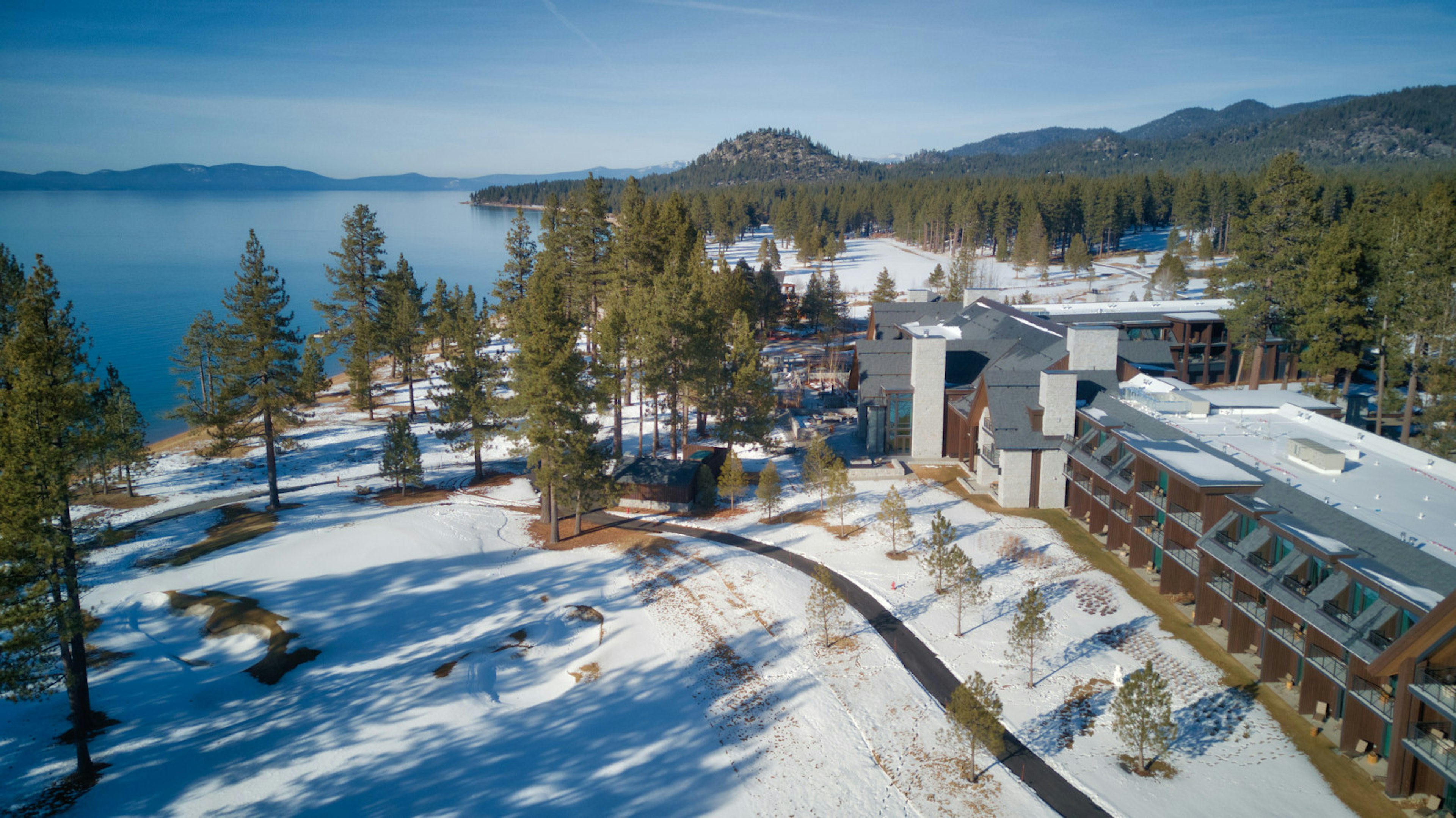 Aerial shot. The ground is covered in snow, there are tall pine trees everywhere, a calm blue lake on the left and an apartment block on the right with mountains behind it.