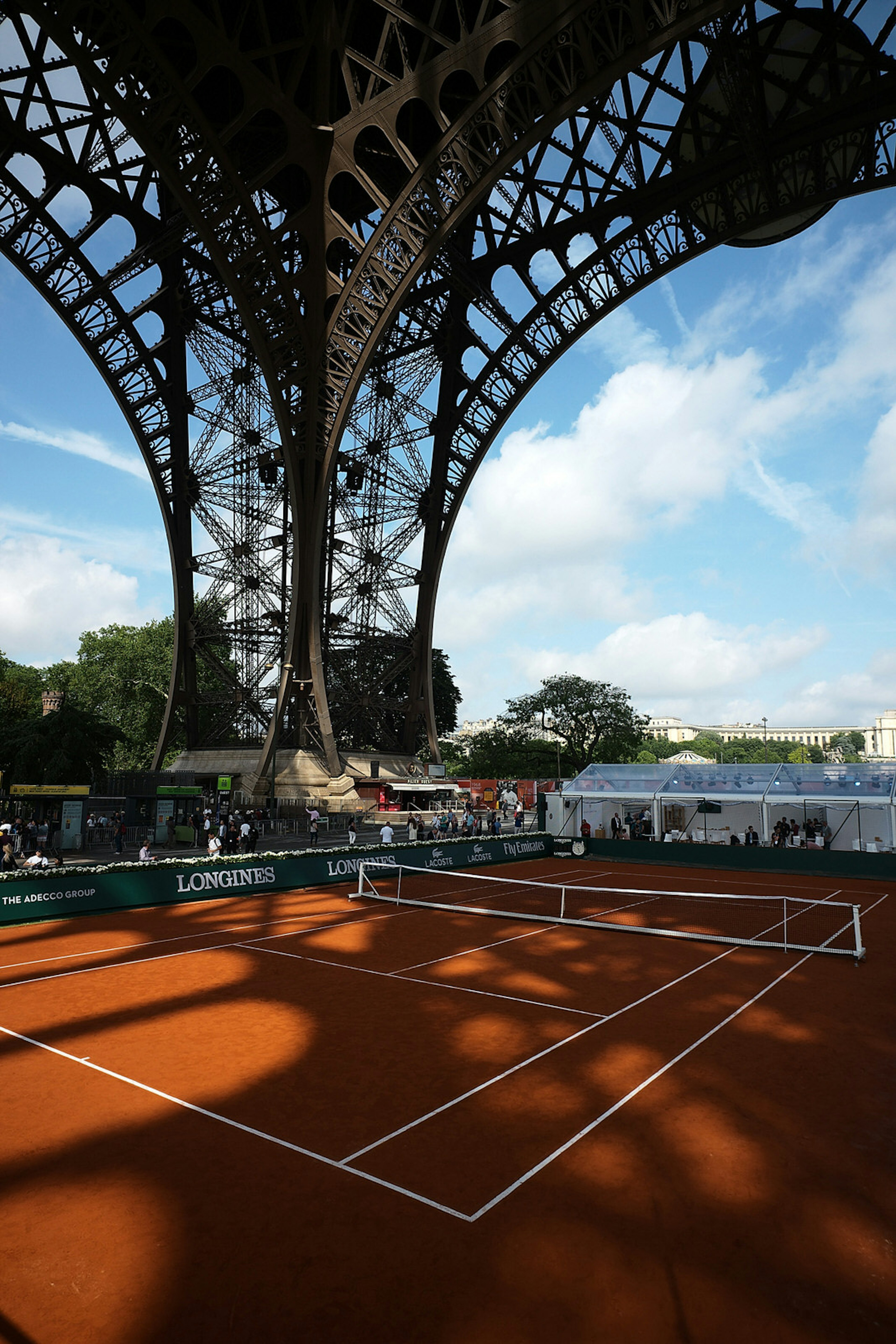 A red clay tennis court, with crisp white lines, sits beneath the elegant iron arches of the Eiffel Tower's large feet; ornate shadows cast by the steel structure spread across the court, with a blue sky in the background