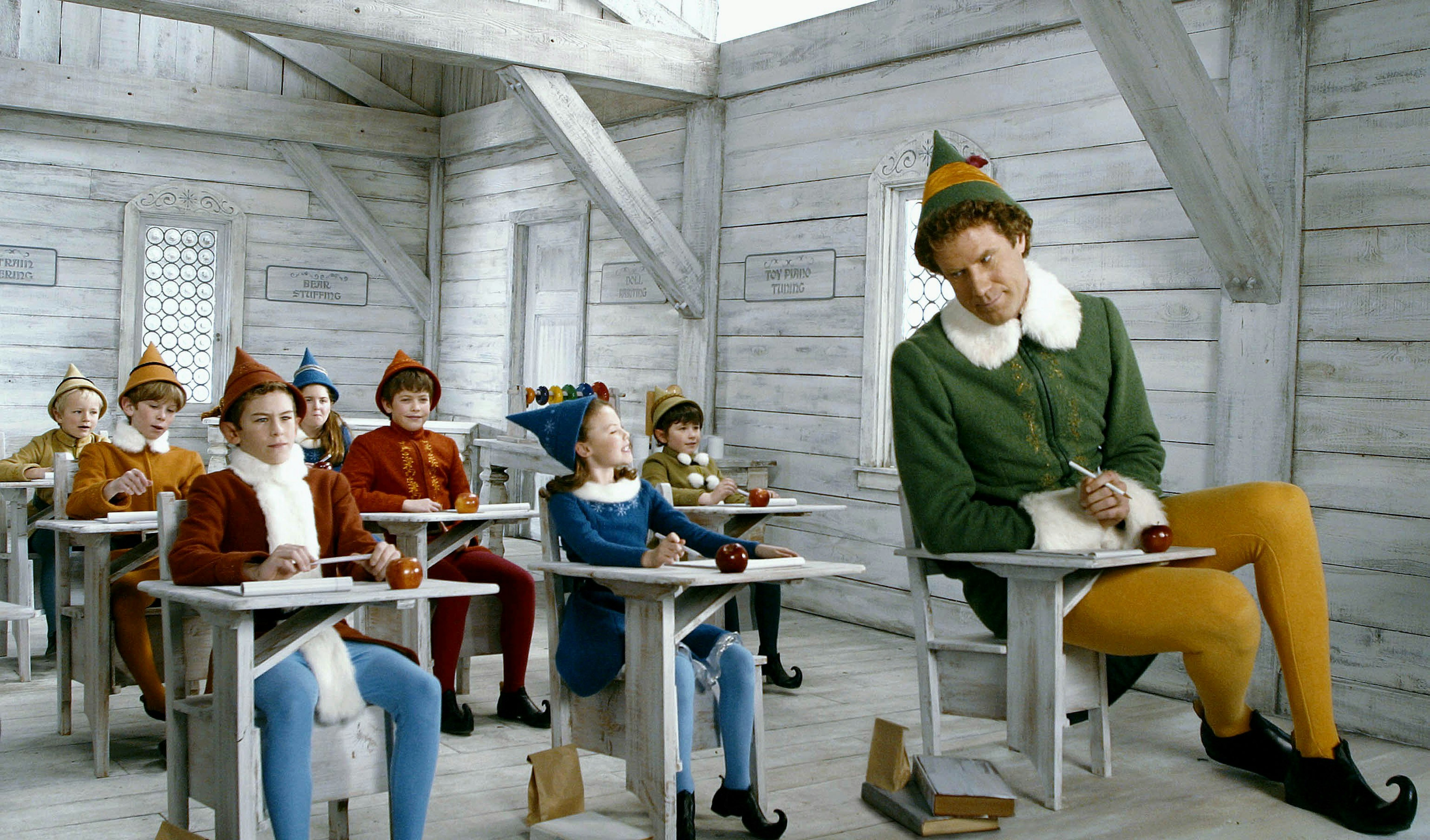 A smirking Will Ferrell, dressed as an elf, sits in a classroom full of elves. He is much bigger than them.