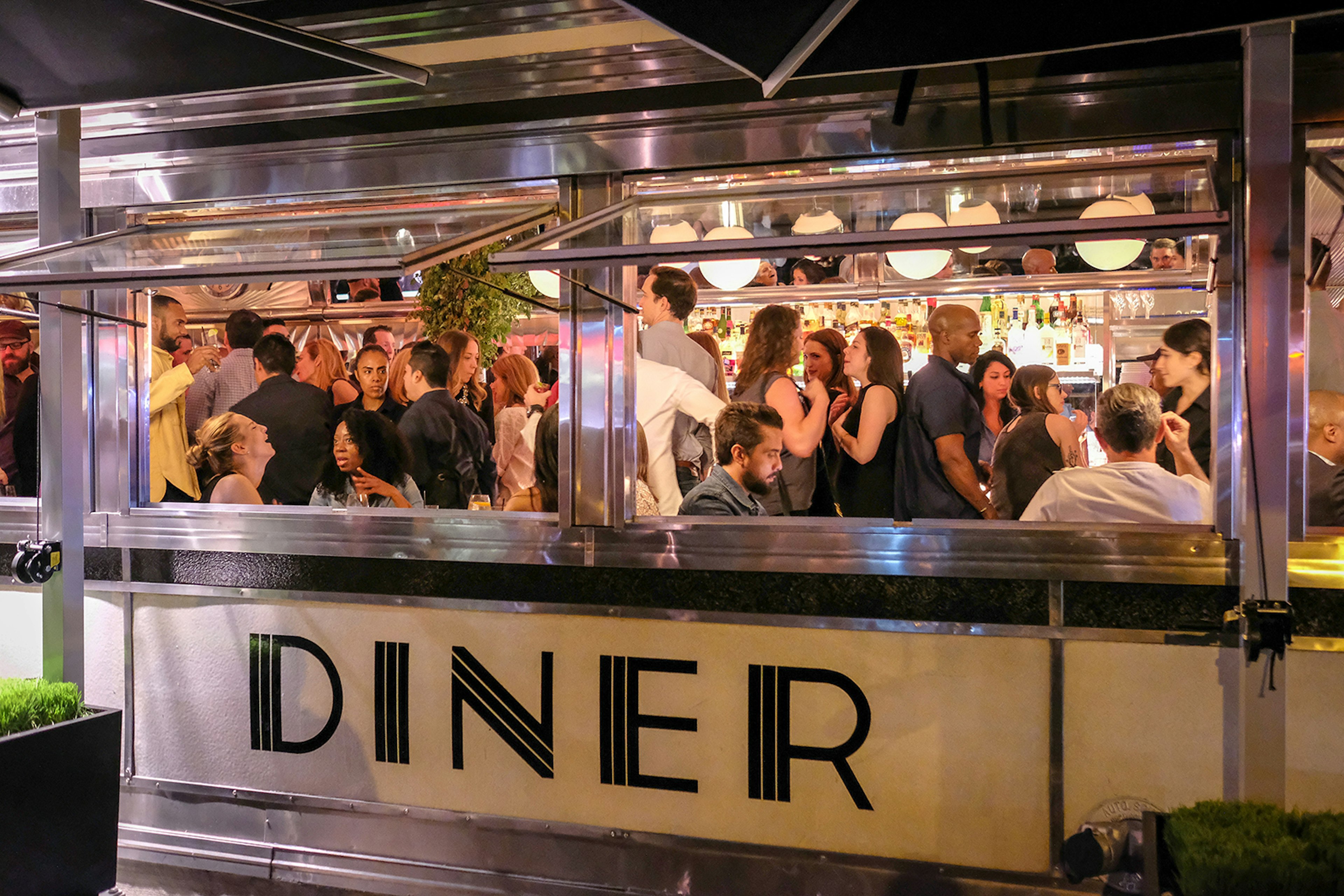 People stand inside a warmly lit New York City diner; its windows are open and the external wall reads