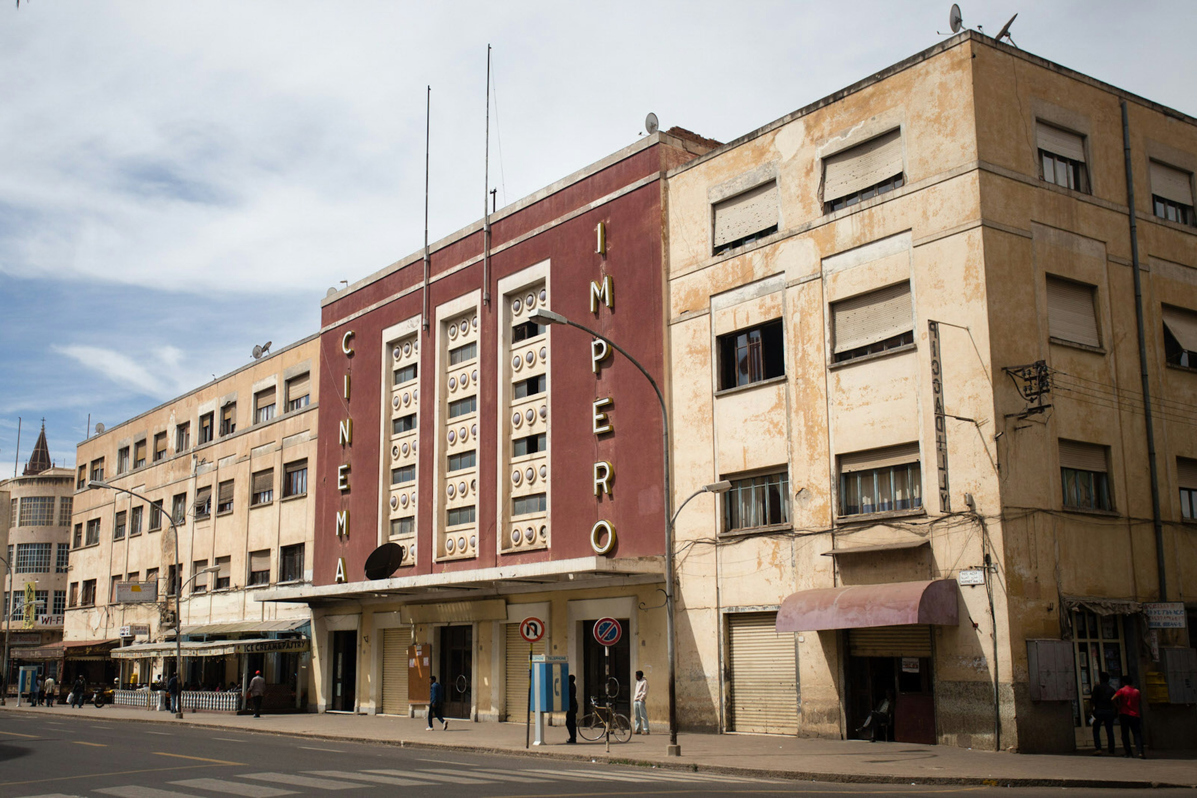 Flanked by two bland, sand-coloured buildings is the red, sleek-looking Cinema Imperio. Above the cantelevered cement awning that runs the length of the building are three symetrical rows of window climbing vertically up the building. On one side of them 'CINEMA' is written vertically in bold gold letters, on the other side is 'IMPERIO'