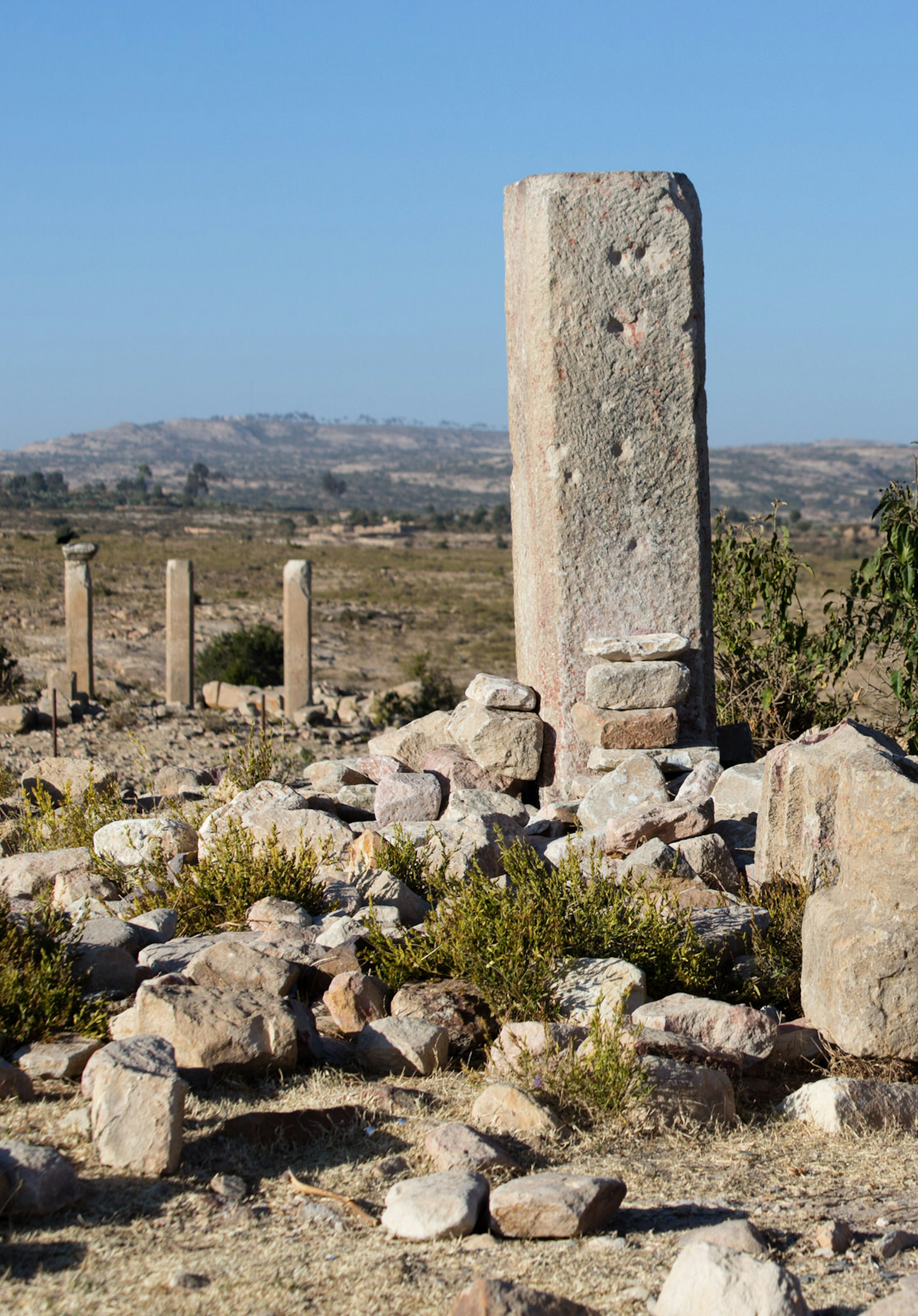 One large stone stella stands tall above a ground strewn with rubble from an ancient Aksumite city in Qohaito, Eritrea. In the background are three smaller stellae and distant rolling hills