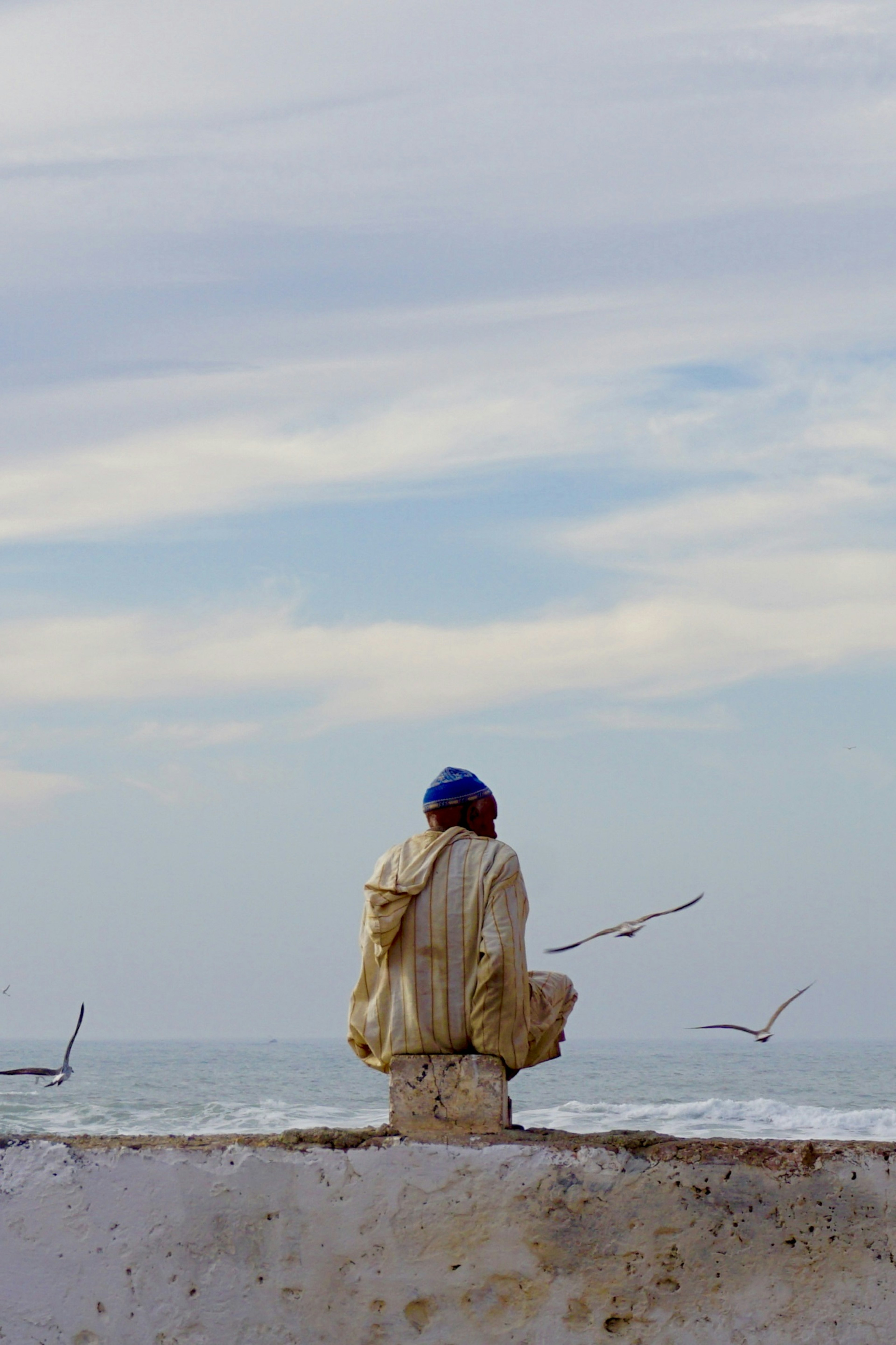 A Berber man sits atop a wall facing the sea (with his back to the camera); the ocean is visible in the distance, and seagulls fly about.