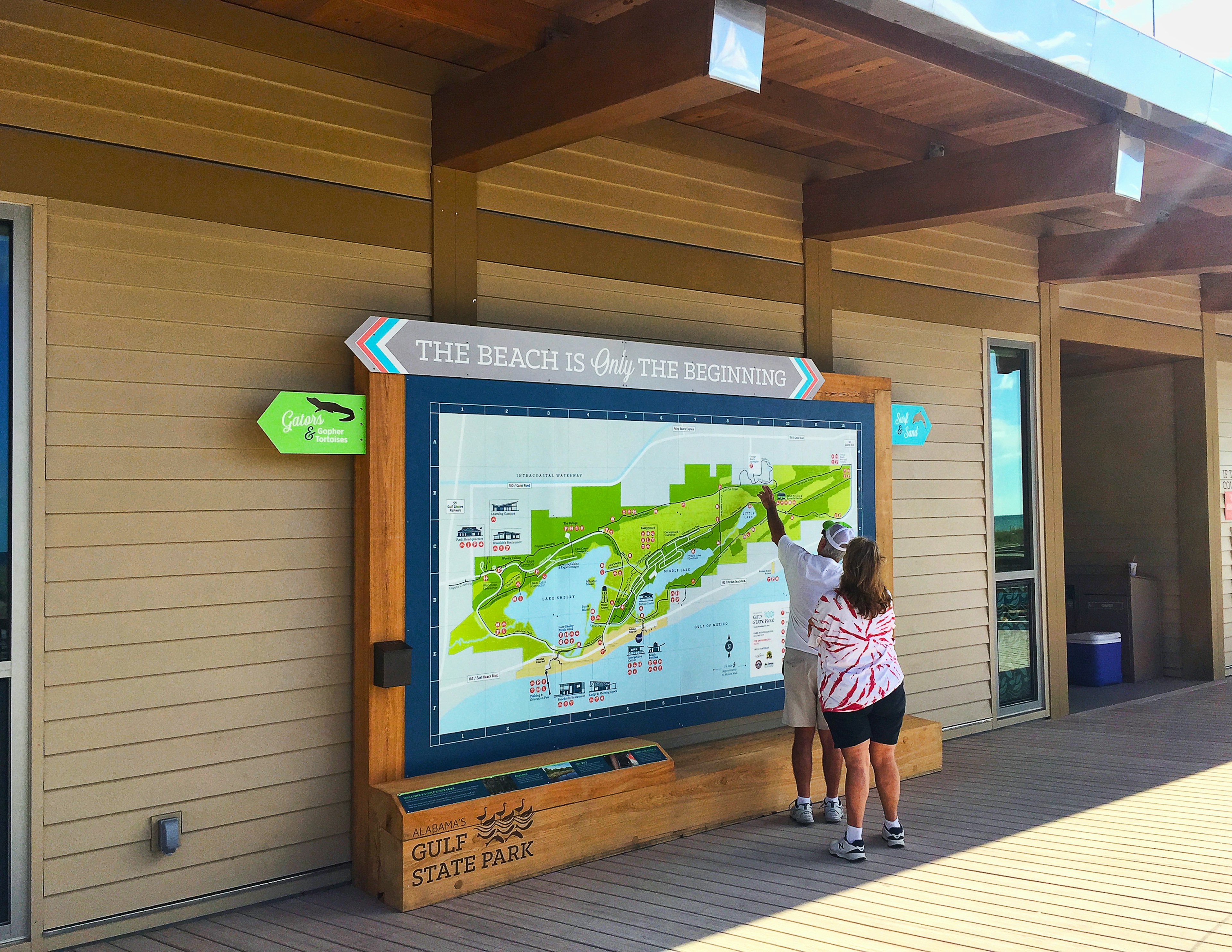 Dressed in shorts and t-shirts, a man and a woman look at a large orientation map of Gulf State Park on wooden decking under a veranda. The man is pointing at something on the map, and a sign above it says