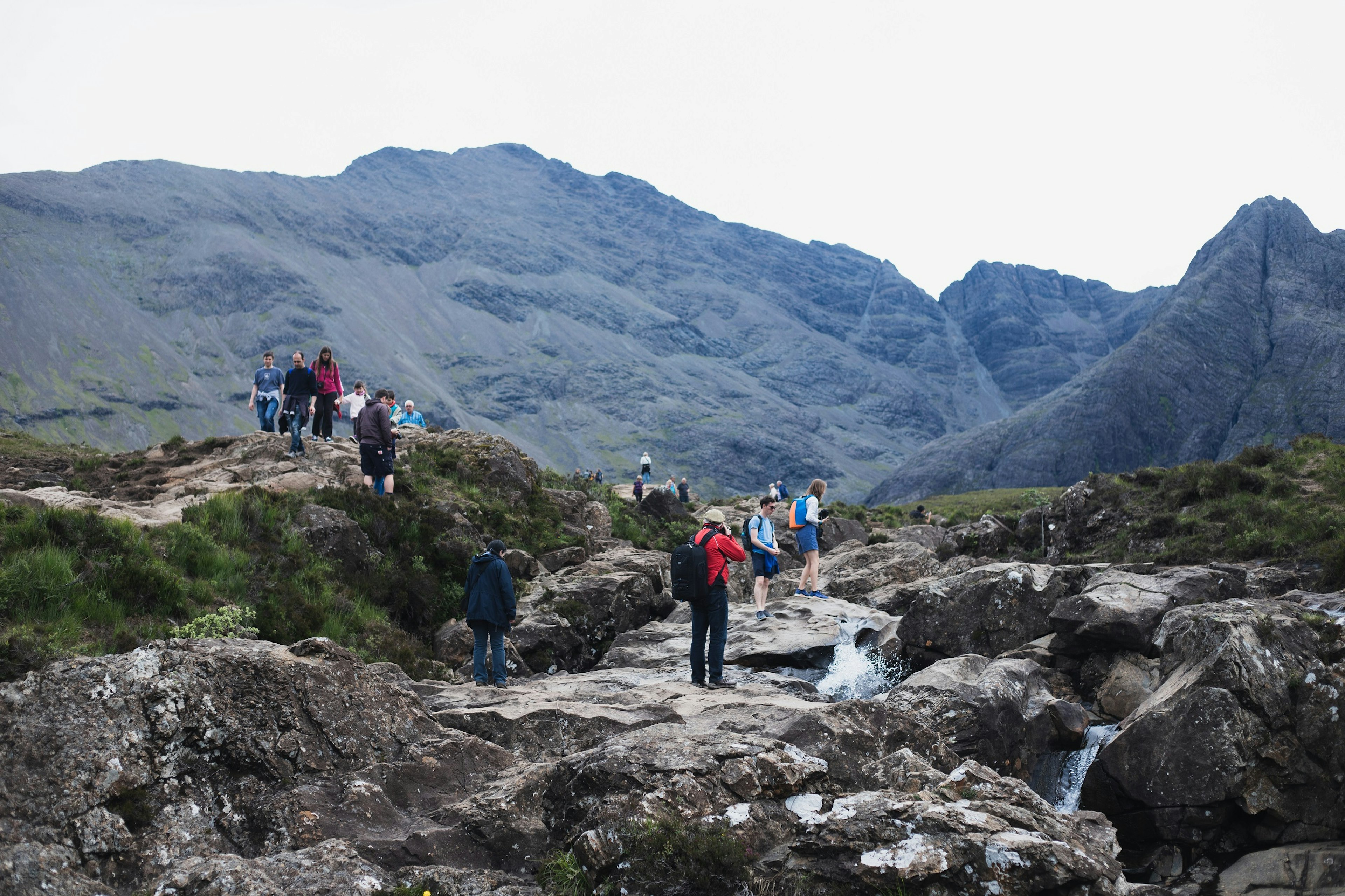 Groups and individuals stand in a rocky area with hills behind and pools of water below. Many are holding cameras.