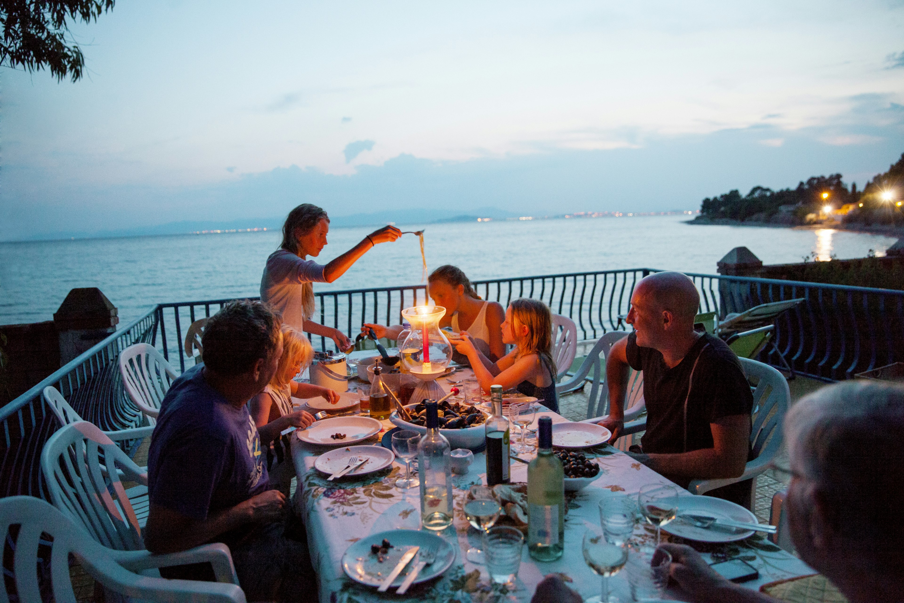 A group of men and women are enjoying a meal outside by the sea at dusk